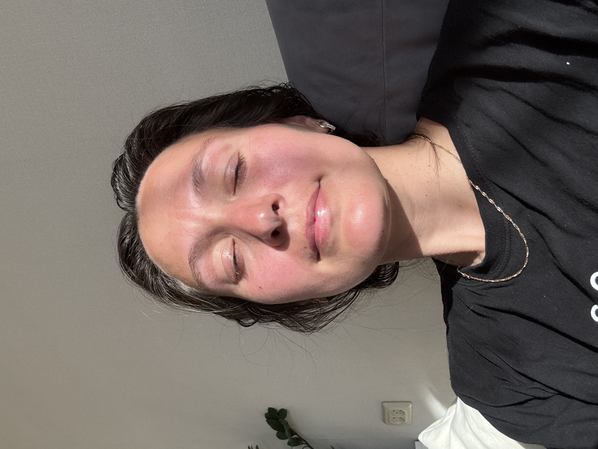 A female individual is captured in a mid-shot, with her eyes closed and head slightly reclined, exhibiting a subtle smile. Her face is illuminated by direct, warm light from the left, resulting in prominent highlights and a flushed, reddish hue across her forehead, cheeks, and nose. Her skin appears dewy or shiny, and her lips are slightly parted and glossy. Her dark hair, possibly damp or slicked back, shows visible strands of gray at the hairline above her left temple. She is wearing a plain black crew-neck t-shirt, and a thin gold-colored chain necklace is visible around her neck. A small silver-toned hoop earring is present in her right ear (viewer's left).
The background to her left features a light-colored wall with a white electrical outlet containing two receptacles. A portion of a green leafy plant is partially visible behind her left shoulder. A white pillow or cushion is also partially visible at the bottom left. To her right, a dark gray or black cushioned surface, likely a pillow or part of a chair, is present. The overall setting appears to be indoors, under strong natural or artificial light.