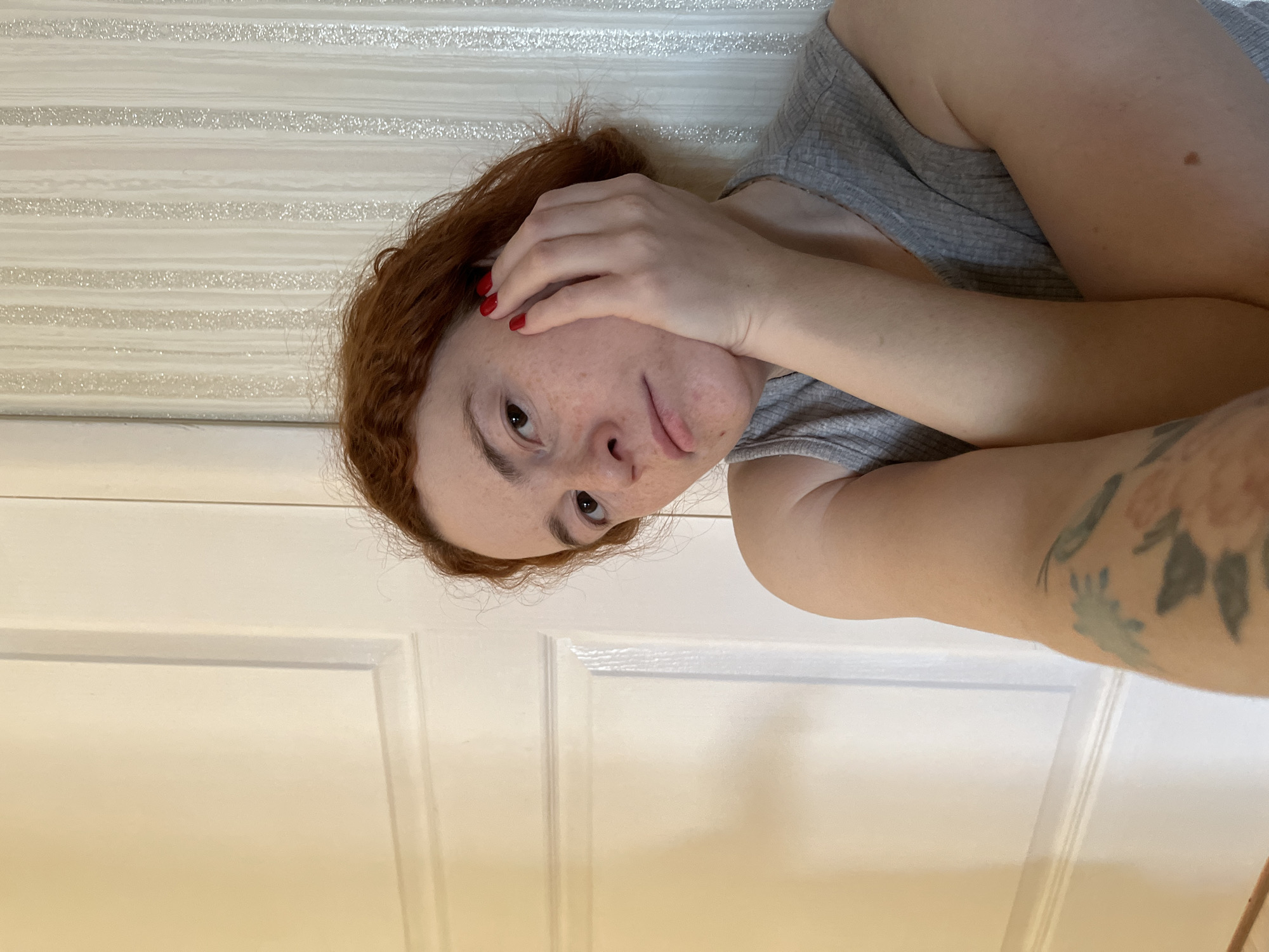 An overhead, full-body shot captures a young individual, likely female, with red or auburn curly hair, lying down in an indoor setting. She is looking towards the camera with her left eye, while her right hand, adorned with red nail polish, covers part of her forehead and right eye. Freckles are visible across her nose and cheeks. Her mouth is slightly open, hinting at a neutral or slightly pensive expression. She wears a grey, ribbed tank top or camisole. A tattoo featuring several muted floral designs and leaves is visible on her left arm.
The background consists of two distinct sections. The upper half features a wall with horizontal stripes, some of which appear to be glittery or textured, reflecting light. The lower half of the image shows a white paneled door, indicative of an interior room. The lighting is soft and even, suggesting either diffused natural light or gentle artificial illumination. There are no strong shadows, making the time of day indeterminate. No specific weather conditions can be inferred from the indoor scene. The provided location context is Chaikovsky, Russia, though no specific visual cues within the image confirm this. There is no visible text in the image.