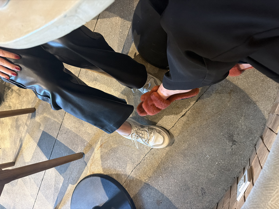 An overhead shot shows two pairs of legs under a table. On the left, someone wears wide-leg black faux leather pants and white sneakers. On the right, another person wears black pants and a pair of fuzzy, reddish-brown bunny slippers. The setting appears to be a café or similar establishment, with a gray concrete floor and a chair visible on the left.
