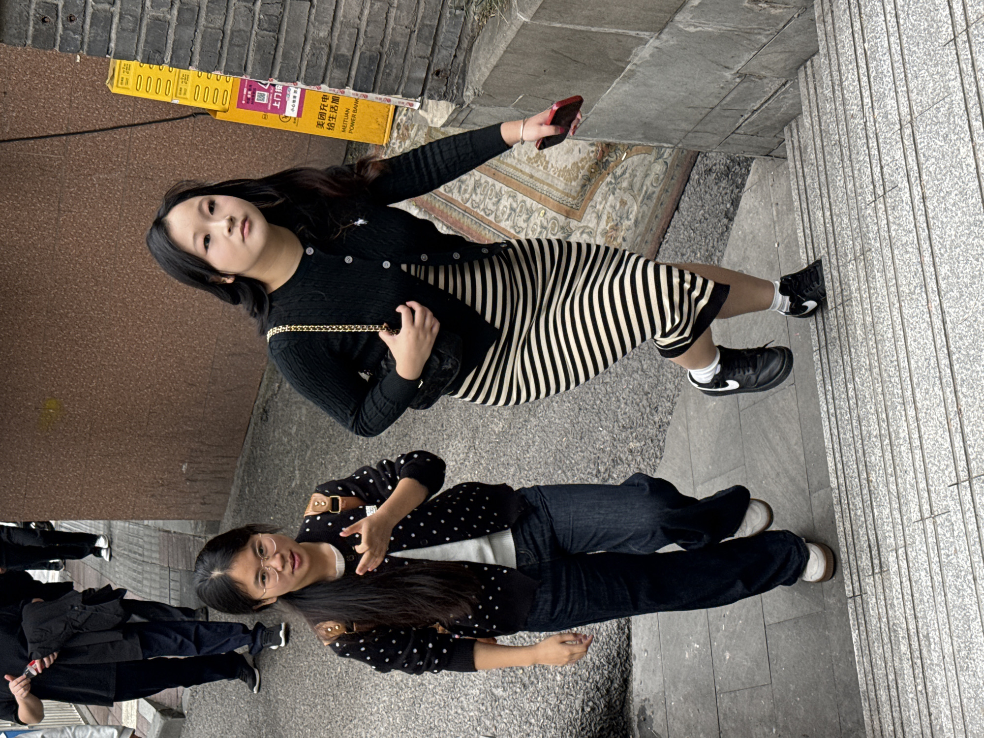 An outdoor scene in Yuzhong District, China, depicts two young East Asian women descending a grey concrete staircase. The woman on the left, positioned slightly behind the other, wears clear-framed glasses, a black cardigan with white polka dots over a white shirt, dark blue wide-leg jeans, and light-colored platform sneakers. She has long dark hair tied back and gestures with her left hand, palm open and fingers bent, while looking directly towards the viewer. Brown leather backpack straps are visible on her shoulders. The woman on the right, in the foreground, wears a black cardigan over a black and white horizontally striped midi dress, black sneakers, and white socks. She holds a red smartphone in her right hand and carries a black shoulder bag with a gold chain strap on her left shoulder, looking slightly upwards and to the left. Behind the women, on a flat paved area, multiple individuals are visible, with one person in a black t-shirt holding a red-capped bottle and a black object. To the right of the staircase, a tall, yellow vertical sign, partially obscured by a dark grey brick wall, displays "美团充电 续生活加" (Meituan Power Bank, life continues) and "MEITUAN POWER BANK" in black text. The wall on the left is a textured brown. An ornate, patterned rug or tapestry lies discarded or integrated into the ground adjacent to the stairs on the right side.