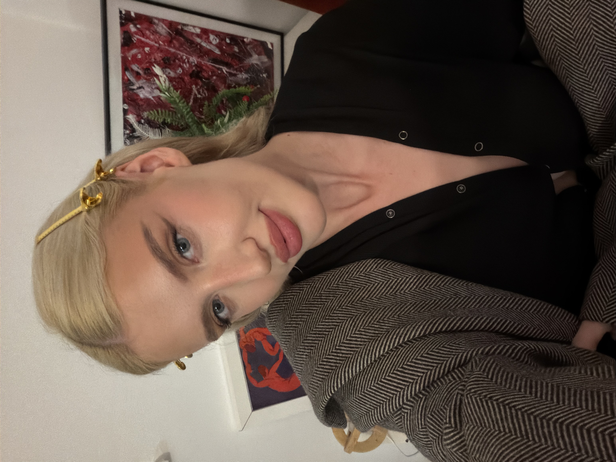 A light-skinned woman with blonde hair is shown in a close-up, tilted-angle portrait. She has fair skin, blue eyes, and is wearing a gold headband. She is wearing a black top with visible snap closures and a brown and white herringbone jacket. Behind her are two framed artworks on a white wall. The specific location in the city of Vozdovac Urban Municipality, Serbia cannot be determined from this image.