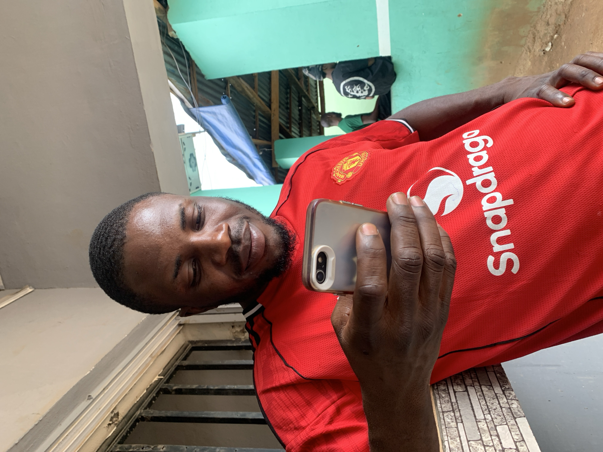 The image, rotated 90 degrees clockwise, captures a man holding and looking at a smartphone. He has dark skin, short dark hair, and a short, dark beard, with a slight smile or smirk on his face. He is wearing a red short-sleeved jersey.
Visible details on his jersey include a yellow and black logo, identified as the Manchester United crest, on the left chest. Prominently displayed across the front of the jersey in white capital letters is the text "Snapdragon". The smartphone he holds appears to be a model with dual cameras on the back.
The setting appears to be an outdoor or semi-outdoor area at the New Stadium, Nigeria, during daylight hours. In the background, there is a light-colored wall to the left. To the right and further back, a vibrant mint green wall or ceiling is visible, beneath which are wooden beams and glimpses of two other people – one in a black t-shirt and another in a green t-shirt. A blue tarp or fabric is also present in the background. In the lower left of the frame, a dark metal grate is partially visible, possibly part of a window or barrier. His right arm rests on a surface with a textured, small rectangular tile pattern. The lighting is bright and natural.