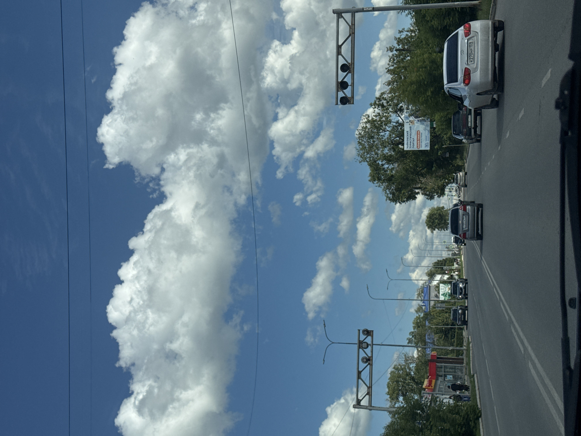 This media file shows a view from a car driving on a road in Chaikovsky, Russia. The scene is a daytime view with a bright blue sky and large, fluffy white clouds. The road appears to be in an urban or suburban area, with trees lining the sides and buildings visible in the distance.
The primary activity depicted is driving, with several cars visible on the road, including a silver sedan in the foreground, a dark SUV behind it, and other vehicles further down the road. Traffic lights are also visible, suggesting an intersection or controlled passage.
Notable details include the presence of power lines running parallel to the road. On the left side of the road, a large billboard is visible with text that appears to be in Russian. One sign reads "ЧАЙКОВСКИЙ ТУРИСТИЧЕСКИЙ ПОРТАЛ" which translates to "Chaikovsky Tourist Portal." The overall impression is of a sunny day, likely during warmer months, and a typical urban street scene. There are no visible signs of distress or unusual events.