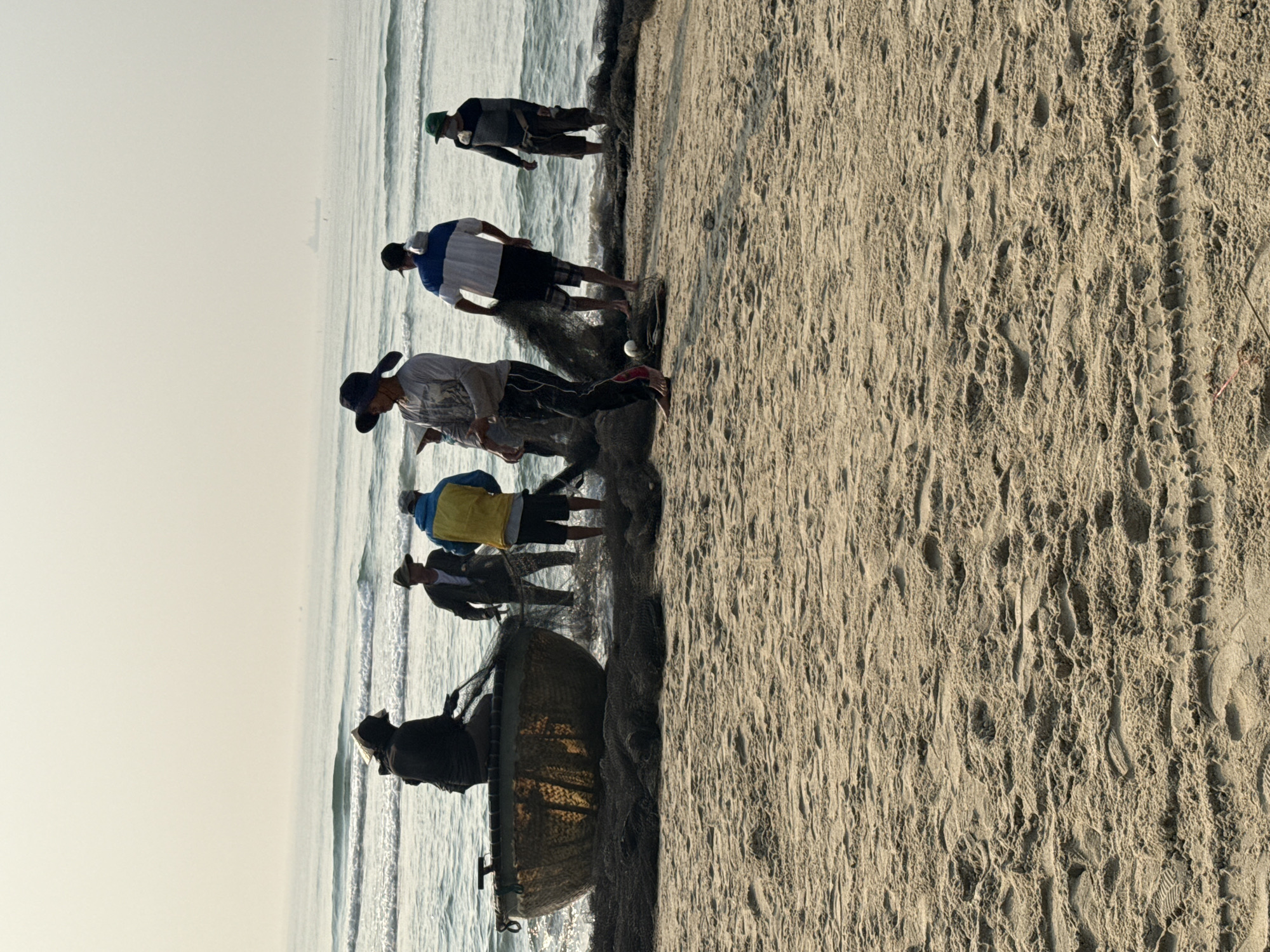 The image shows a group of men fishing on a beach in Đà Nẵng, Vietnam. The scene is divided between the sandy beach on the right and the ocean on the left. Six men are visible at the edge of the water, interacting with a large fishing net. One man is holding a round boat-like structure. The sky is hazy. The beach shows various patterns, suggesting human activity and possibly tire tracks. The waves are small and consistent.