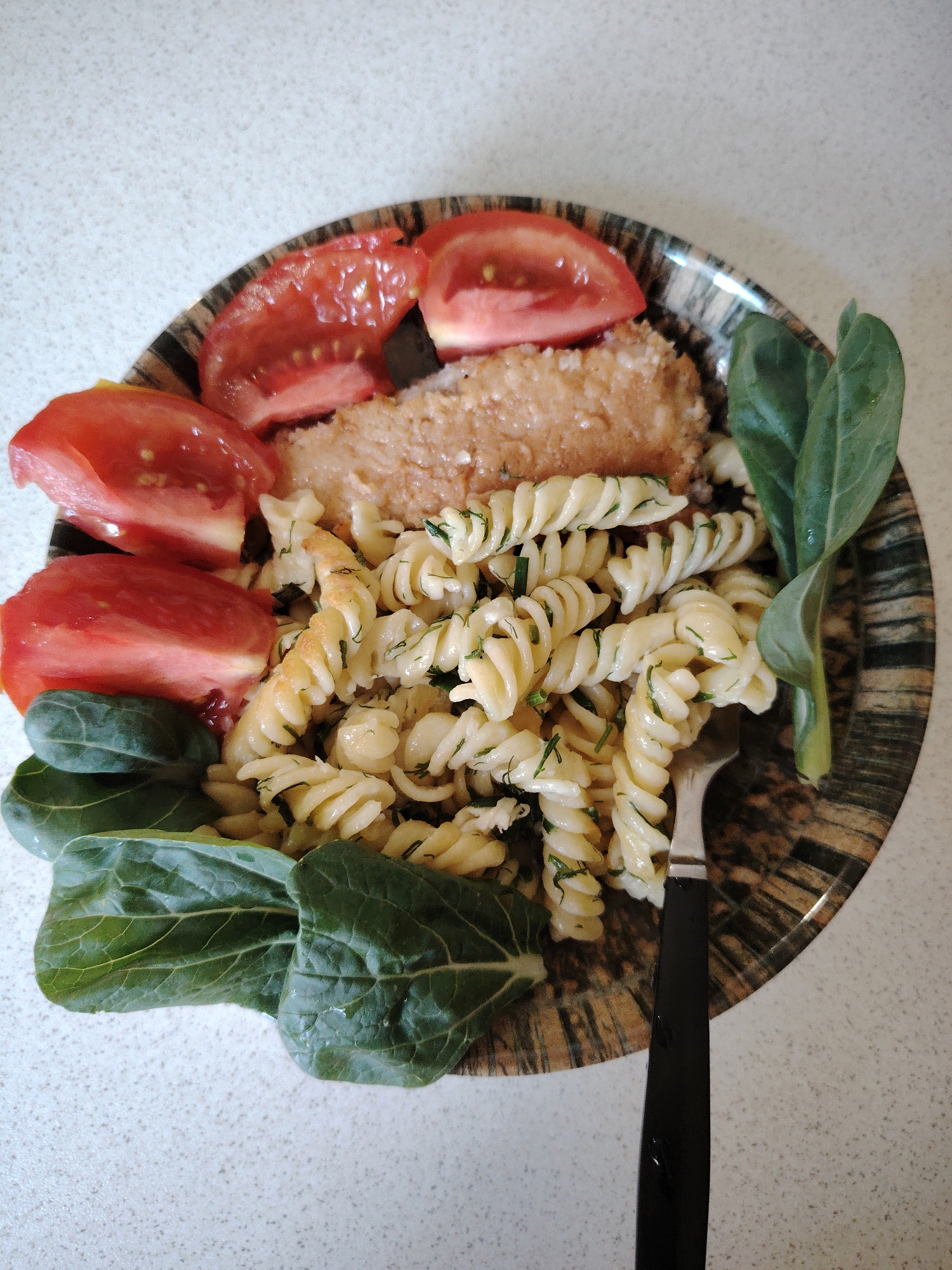 A close-up, overhead shot shows a plate of food. The plate is round with a dark, wood-grain pattern. In the center of the plate is a portion of fusilli pasta with chopped green herbs mixed in. To the left of the pasta are three tomato wedges, and behind the pasta is a light brown, oblong piece of food, possibly a chicken cutlet or fish fillet, breaded and cooked. Several large, dark green spinach leaves are arranged around the edge of the plate. A fork with a black handle rests on the right side of the plate, partially submerged in the pasta. The plate is on a speckled, off-white countertop. The scene is indoors, with no visible windows or doors. The lighting suggests it is daytime. The food appears to be a complete meal. The image contains no people or any indication of the city of Vladimirovka, Russia.