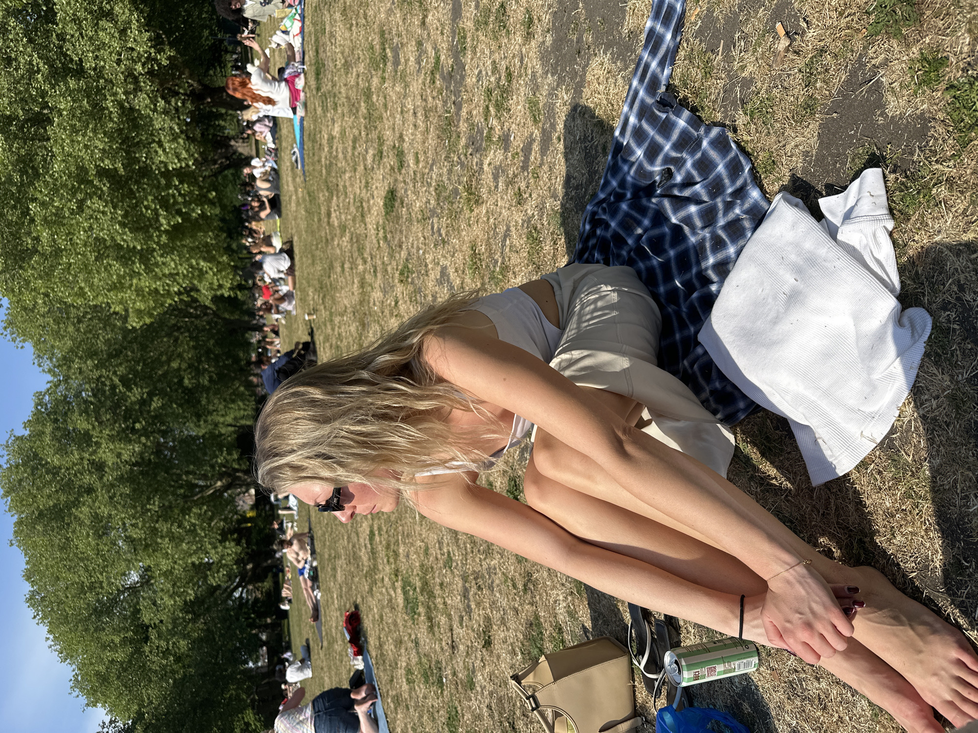 A sunlit, eye-level, medium shot shows a young woman sitting on dry grass, with a crowd of people sitting in the background.
The woman is fair-skinned with long blonde hair, wearing a white top, and light colored shorts. She is wearing dark sunglasses. Her legs are crossed in front of her and her bare feet are visible. A gold bracelet is visible on her right wrist and a black bracelet is on her left wrist. Her hands are clasped together.
A dark blue and white plaid blanket is beside her, topped with a white sweatshirt. Below her legs is a brown purse, sunglasses, a blue bag, and a green and white can.
In the background, a crowd of people are sitting on the grass, shaded by green trees. The sky is pale blue.
Given the crowd of people in a grassy area, with trees, the location is likely a park in London, United Kingdom.