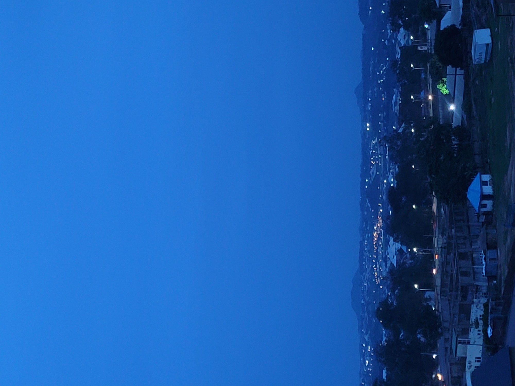 This is a nighttime aerial view of Gura Top, Nigeria. The image shows a sprawling cityscape illuminated by streetlights and building lights. In the foreground, there are several buildings under construction, with exposed concrete structures and scaffolding. A small blue-roofed house is visible, as well as a white container unit. Further into the city, clusters of houses and larger buildings are discernible, all lit up against the dark landscape. In the distance, rolling hills or mountains are silhouetted against the faint glow of the sky, with a more pronounced peak visible in the center-left. The overall impression is of a developing urban area at dusk or night.