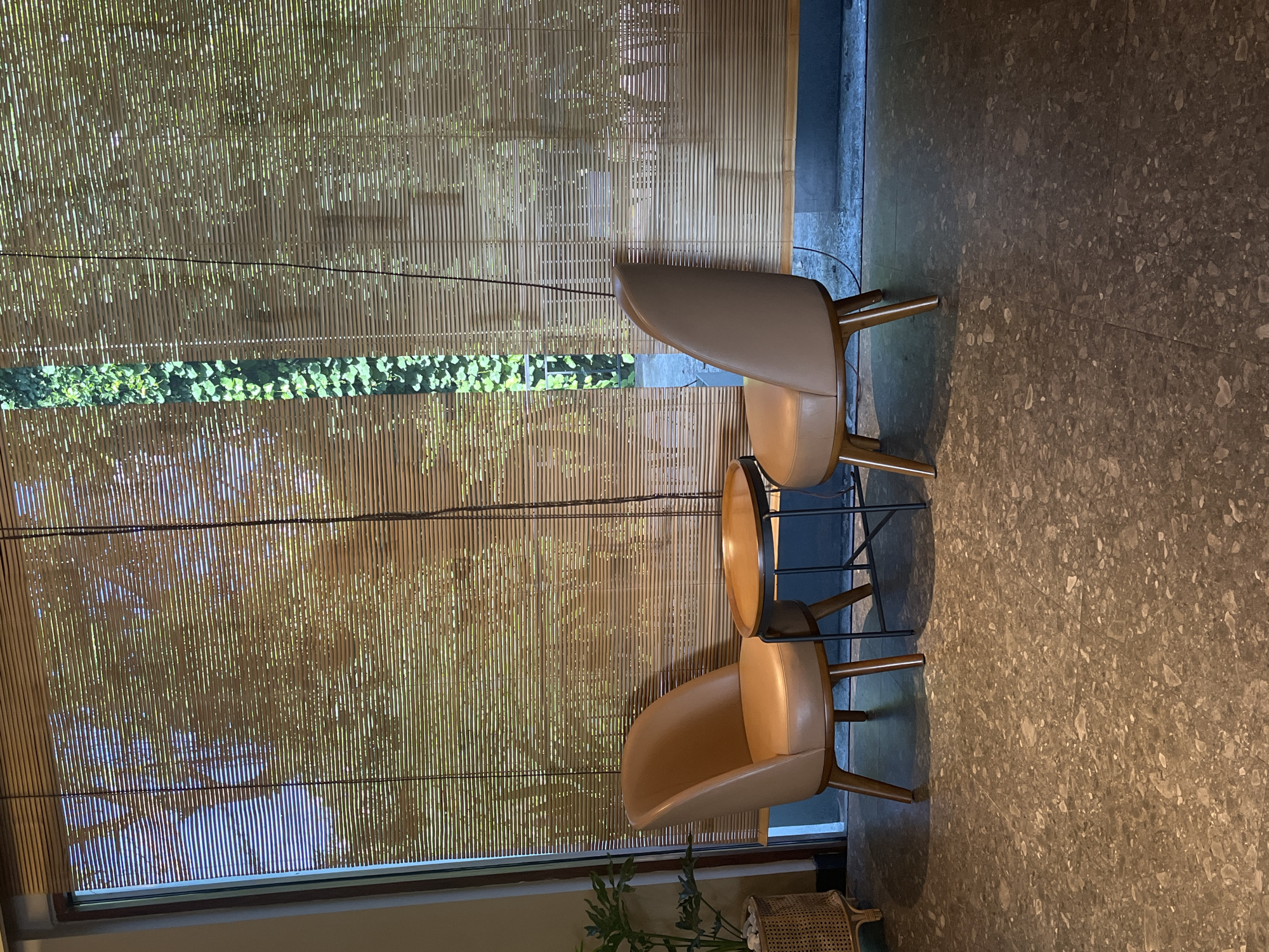 Two modern, light brown leather chairs are positioned facing each other across a small, round wooden table with a dark metal base. The chairs have wooden legs. The scene is indoors, with a window covered by bamboo blinds that allow dappled light to filter through, casting shadows of foliage onto the blinds. To the left, a portion of a large potted plant in a woven basket is visible. The floor is a dark, speckled terrazzo. The setting appears to be a cafe or seating area in a commercial establishment.