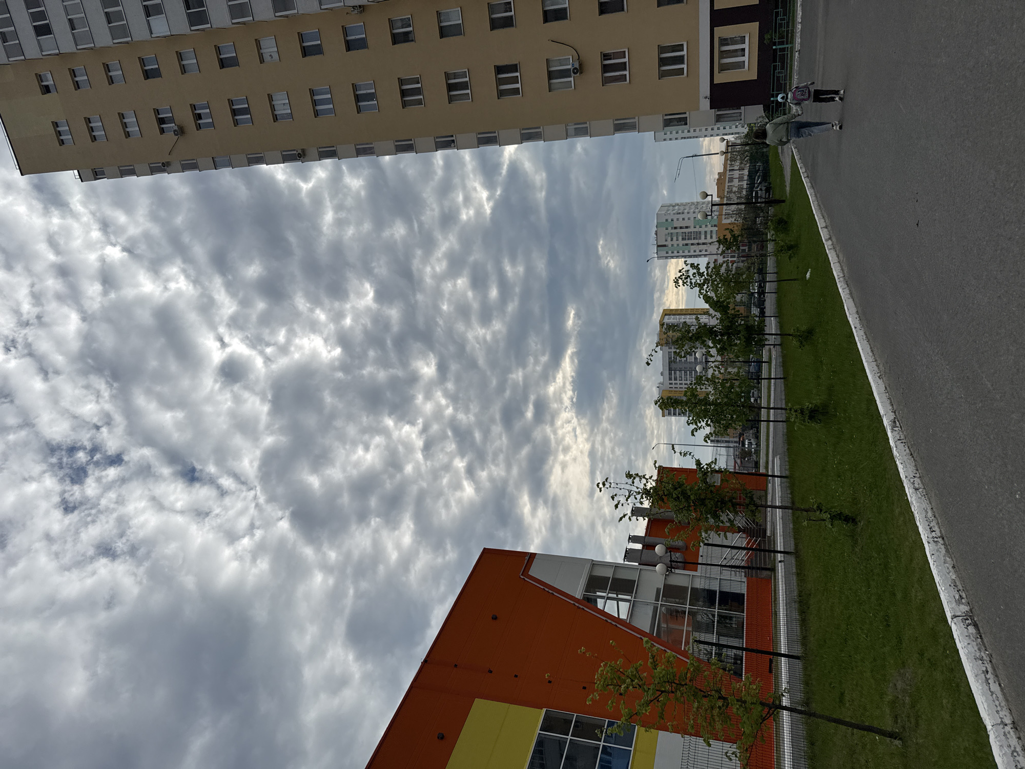A low-angle shot in Tobolsk, Russia, shows an urban landscape. A tall, tan apartment building stands parallel to an orange and yellow commercial building, framing a cloudy sky. Two people walk on the sidewalk. A grassy median with trees separates the sidewalk from the buildings. Other apartment buildings are visible in the distance.