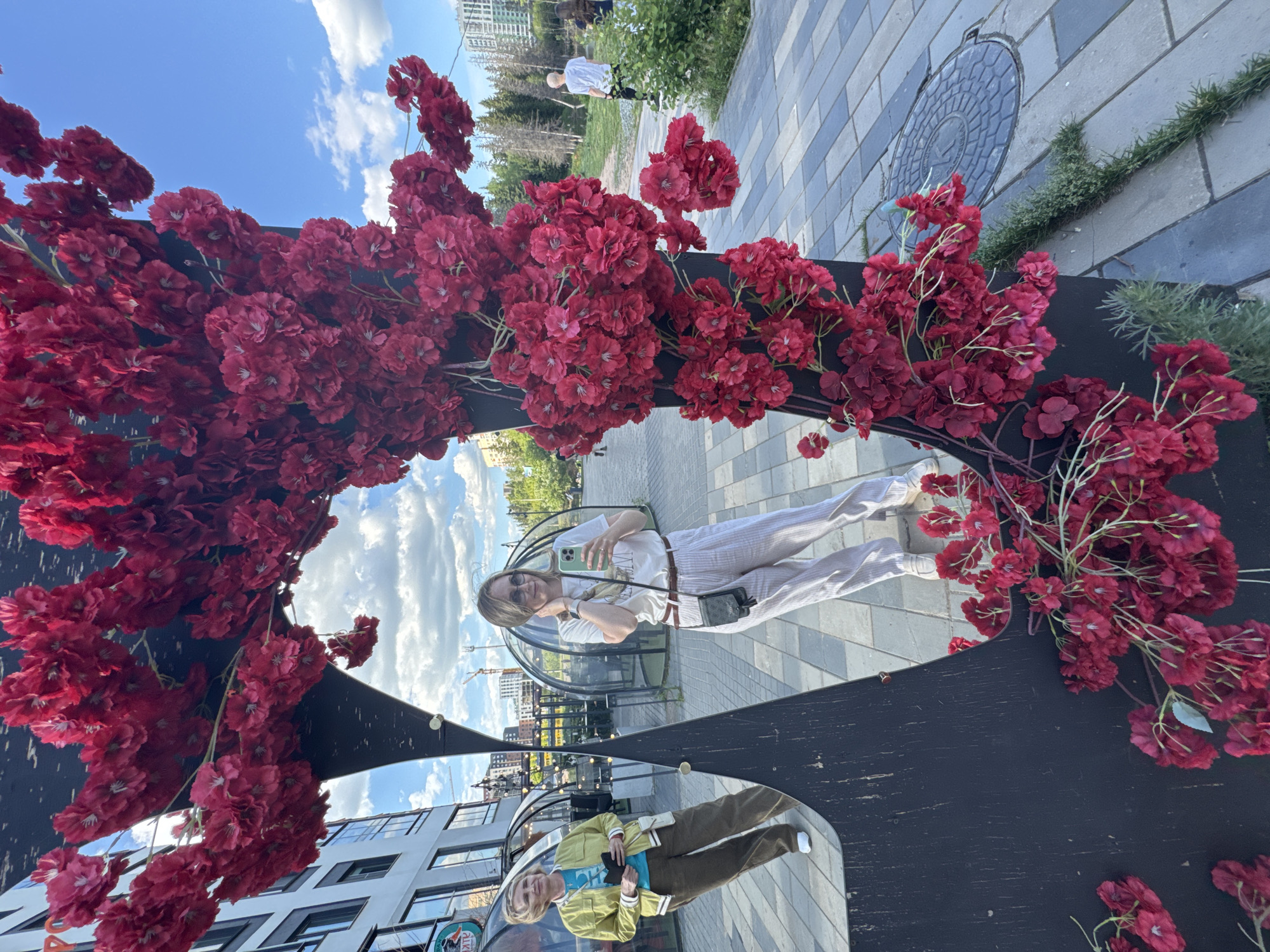 The image captures an outdoor urban scene featuring a large, black, irregularly-shaped art installation covered with dense clusters of artificial red cherry blossoms. The installation incorporates a mirrored surface reflecting two women and parts of the surrounding environment.
On the right side of the mirror, a woman with light-colored hair, wearing sunglasses, a white T-shirt with a bird graphic, and light-colored striped pants, is holding a light green smartphone to her face, appearing to take a selfie. She wears a small dark crossbody bag.
On the left side of the mirror, an older woman with light-colored hair, wearing a yellow jacket over a blue top, dark pants, and white sneakers, stands facing right, holding a dark rectangular object.
The mirrored reflection shows modern multi-story buildings, including one with a partial "КИT" (KITE) sign, and several transparent geodesic dome structures, likely outdoor dining pods. The sky in the reflection is blue with scattered white clouds.
Outside the mirror, the immediate environment consists of a paved walkway made of rectangular grey tiles. To the right, a grassy area with trees and bushes extends into the background, where a person in a white T-shirt is visible walking away, accompanied by another partially obscured individual. A circular metal manhole cover is present on the pavement near the base of the installation. The sky outside the mirror is also clear blue with white clouds.
The location is **Izhevsk, Russia**, specifically identified as the area around the Izhevsk Pond embankment, likely in the vicinity of Gastroport or Leto-Park, due to the distinctive outdoor dining pods and the architectural style reflected.