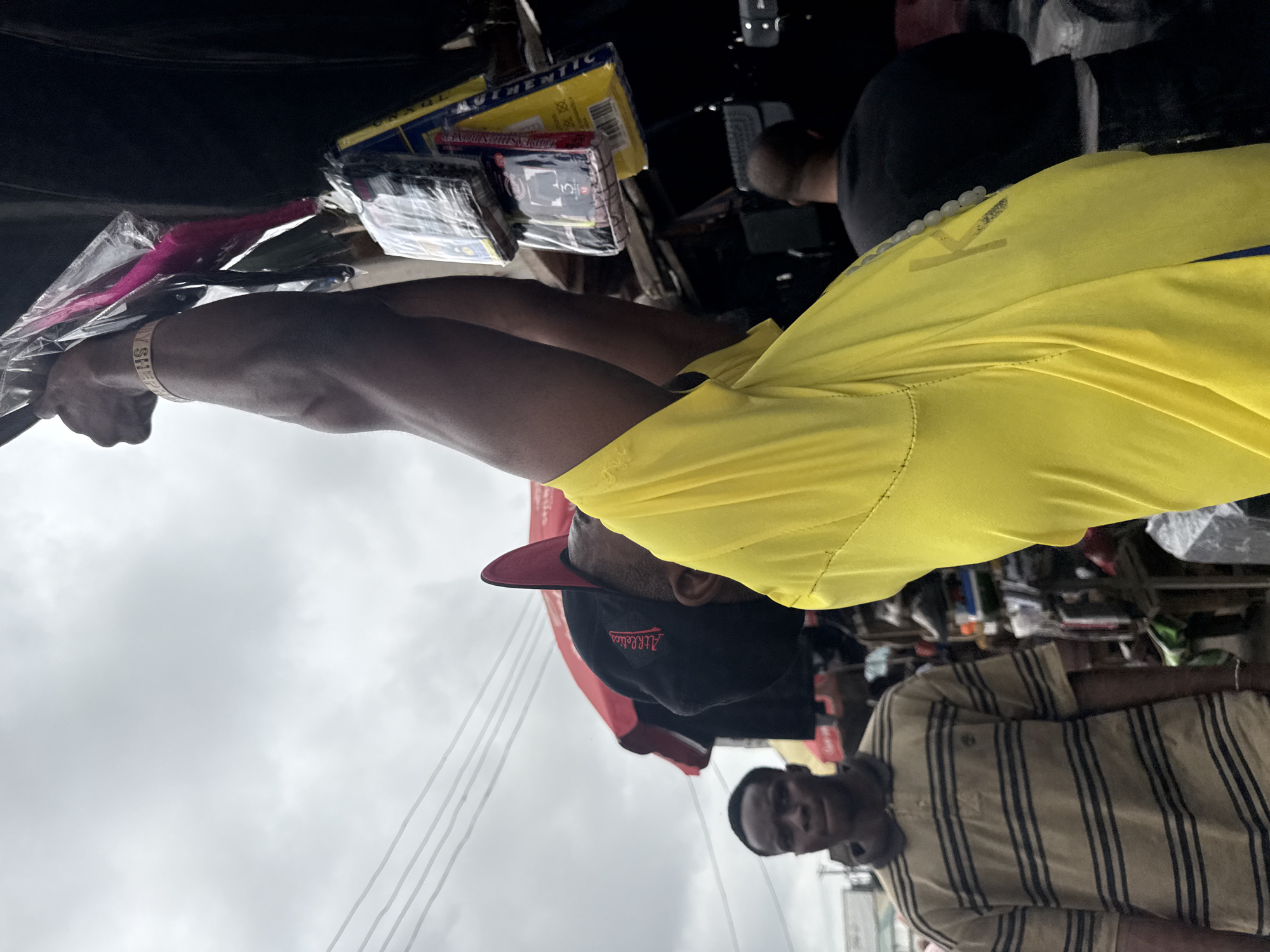 An over-the-shoulder shot captures a Black man in a bright yellow t-shirt raising his arms to adjust merchandise hanging from a stall in an outdoor market. He is wearing a wristband that reads "MY SHEPP." In the background, a Black man wearing a striped polo shirt looks toward the camera. Another Black man in a black shirt is visible further in the background. The market appears to be in a congested urban area, with overhead wires and a red umbrella visible. The sky is overcast. The scene is consistent with an outdoor market stall in Lekki, Nigeria.