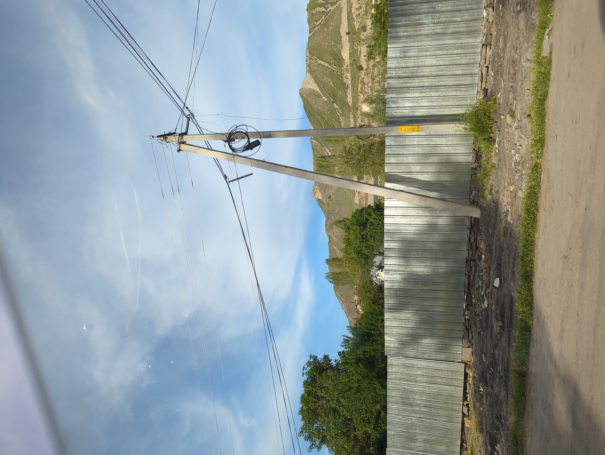 The scene is in Beş-Küŋgöy, Kyrgyzstan. A tilted view shows a weathered utility pole with numerous wires extending to the left and above, set against a blue sky with some clouds. To the right, a corrugated metal fence lines the edge of a paved road with a strip of grass. A rocky embankment with sparse vegetation separates the fence from a mountain visible in the background. Trees are interspersed between the mountain and the fence. A yellow warning sign is affixed to the utility pole near the fence. The perspective is taken from within a vehicle, as suggested by a partial view of the vehicle's interior in the lower left corner.