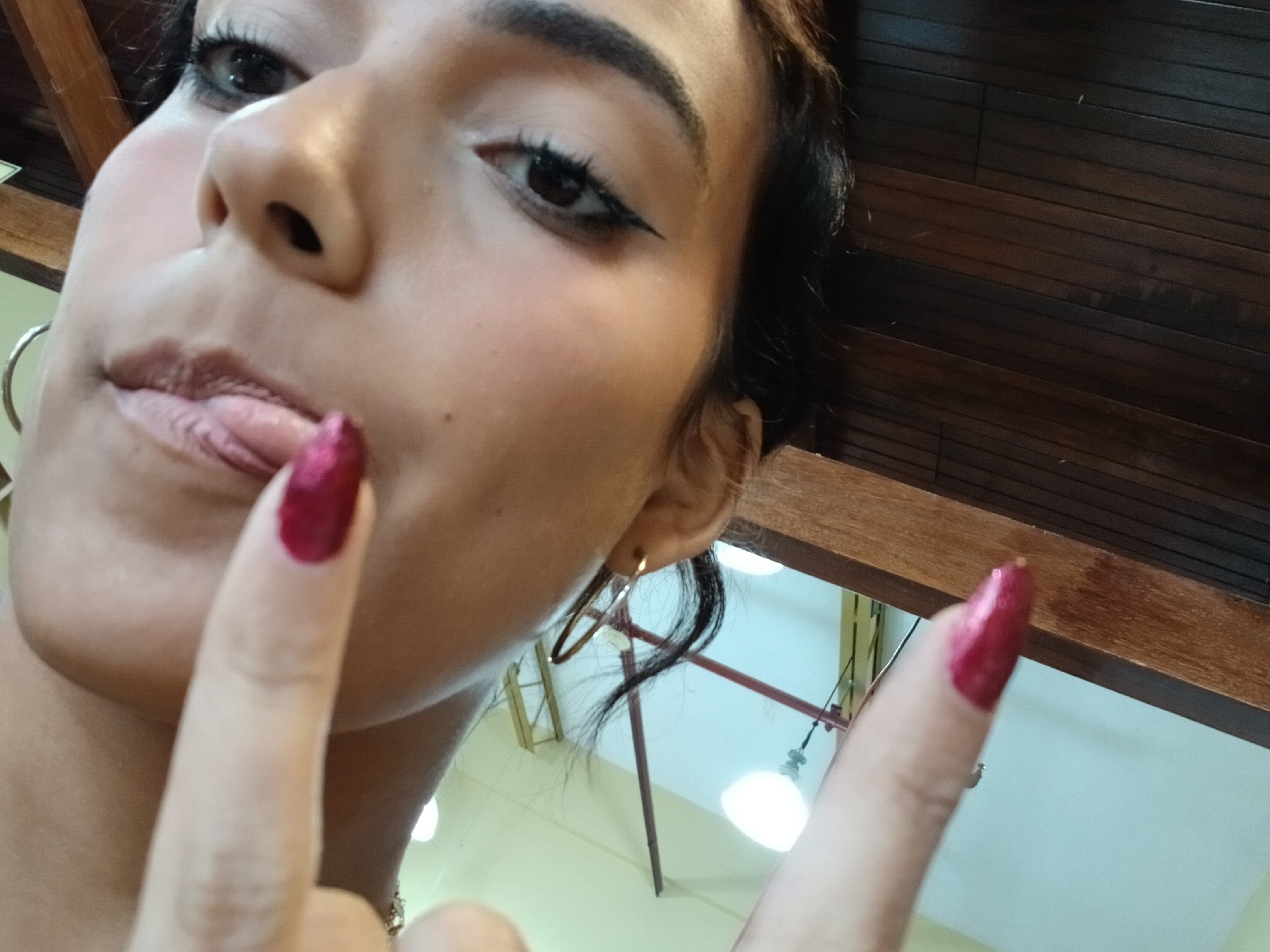 A close-up, low-angle selfie features a person, likely female, looking upwards towards the camera. The individual has a medium skin tone, dark hair, and defined dark eyebrows. Her left eye is visible, accentuated with dark eyeliner and long lashes, gazing directly out of the frame. Her nose and upper lip are prominent in the foreground. Her lips are slightly parted, with her tongue playfully touching her lower lip. A simple, gold-colored hoop earring is visible in her right ear.
Her right hand is raised into the frame, with the index and middle fingers extended. Both fingers display bright, metallic red nail polish. This gesture, potentially a peace sign or a playful signal, adds to the casual and confident demeanor conveyed by the subject.
The background indicates an indoor setting. Dark wooden beams run horizontally across the upper portion of the frame, suggesting a ceiling structure. Below these beams, a lighter-colored ceiling or wall is visible, along with blurred light sources, implying artificial lighting. A reddish-brown metal bar or frame is also faintly discernible in the background.
The image captures a playful and self-assured moment in what appears to be a casual indoor environment, consistent with a location such as Juan Griego, Venezuela. No text is visible in the image.