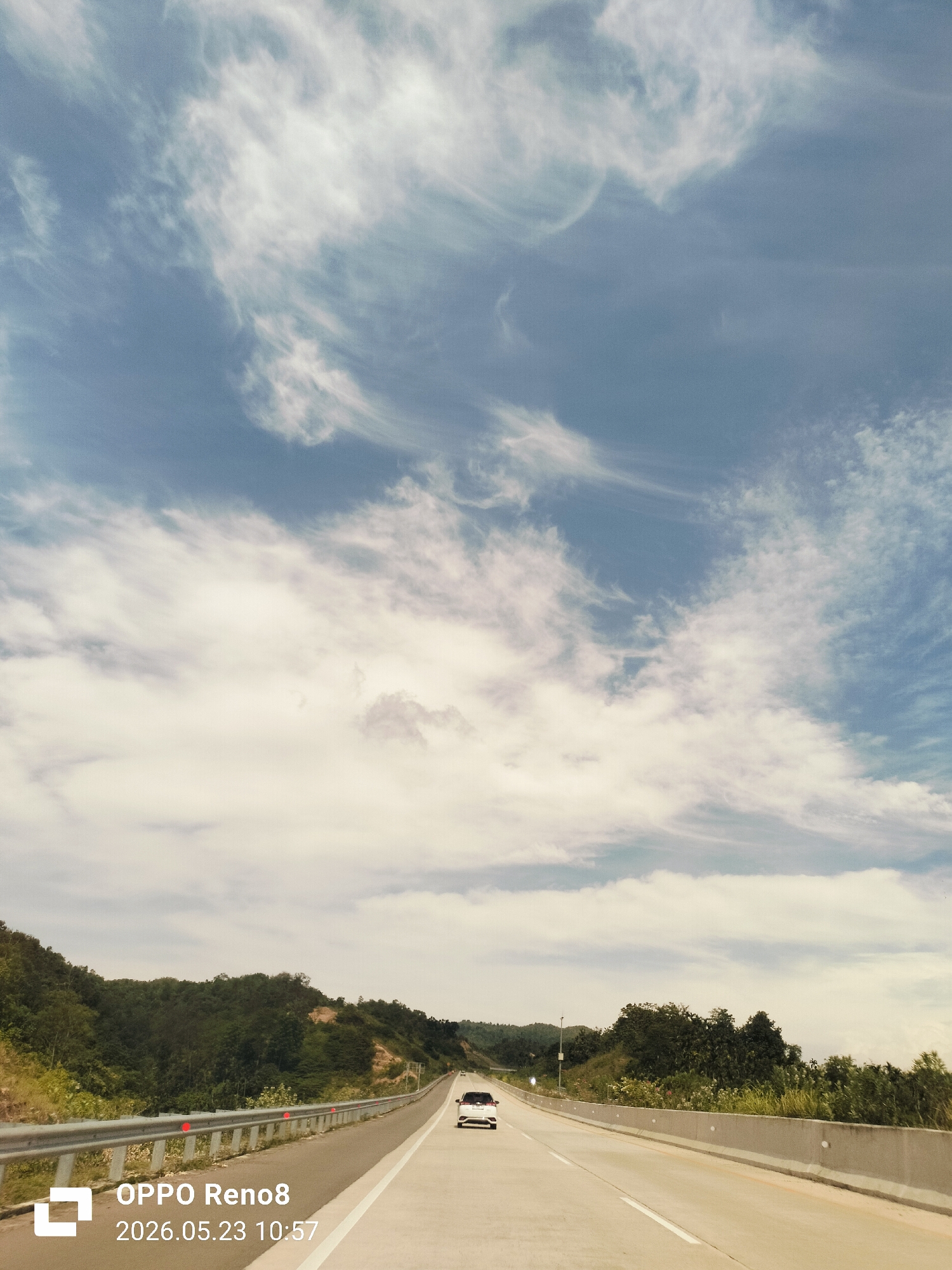 The image depicts a highway in Kreet Paloh, Indonesia, on May 23, 2026, at 10:57 AM. Two cars are visible on the road, traveling in the same direction. The highway is bordered by green vegetation and trees on both sides. The sky is partially cloudy with a mix of blue and white clouds. A gray metal guardrail runs along the left side of the road. The photo was taken with an OPPO Reno8.