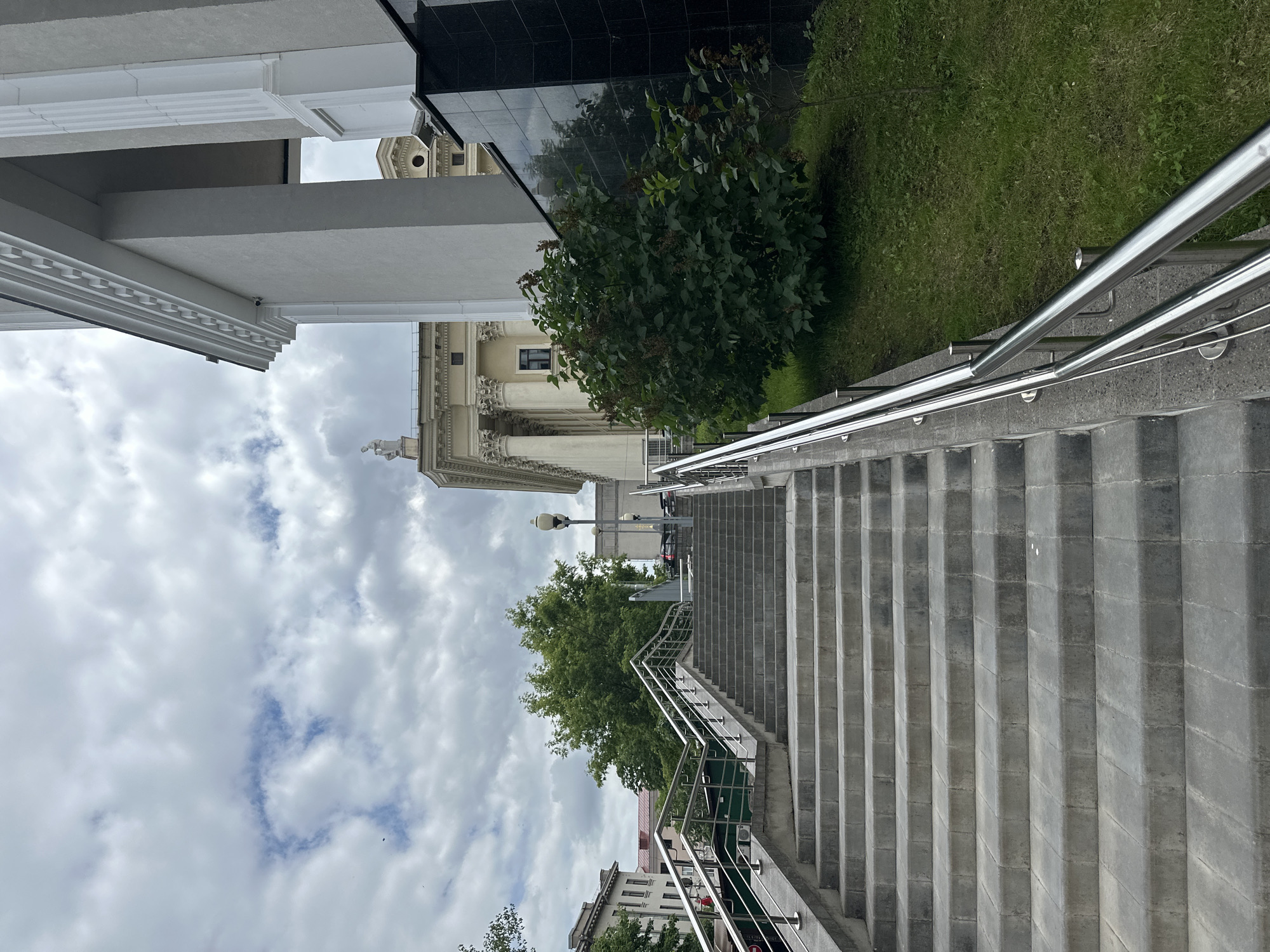 The image shows an outdoor view of a stone staircase leading upwards, flanked by metal railings. The staircase is wide and appears to be made of concrete blocks. To the right of the staircase, there is a lush green bush and a grassy area. In the background, a large building with classical architecture, featuring columns and a statue on its roof, is visible. Further in the background, there are more buildings and trees under a cloudy sky. The location is the National Library of Belarus in Minsk, Belarus.