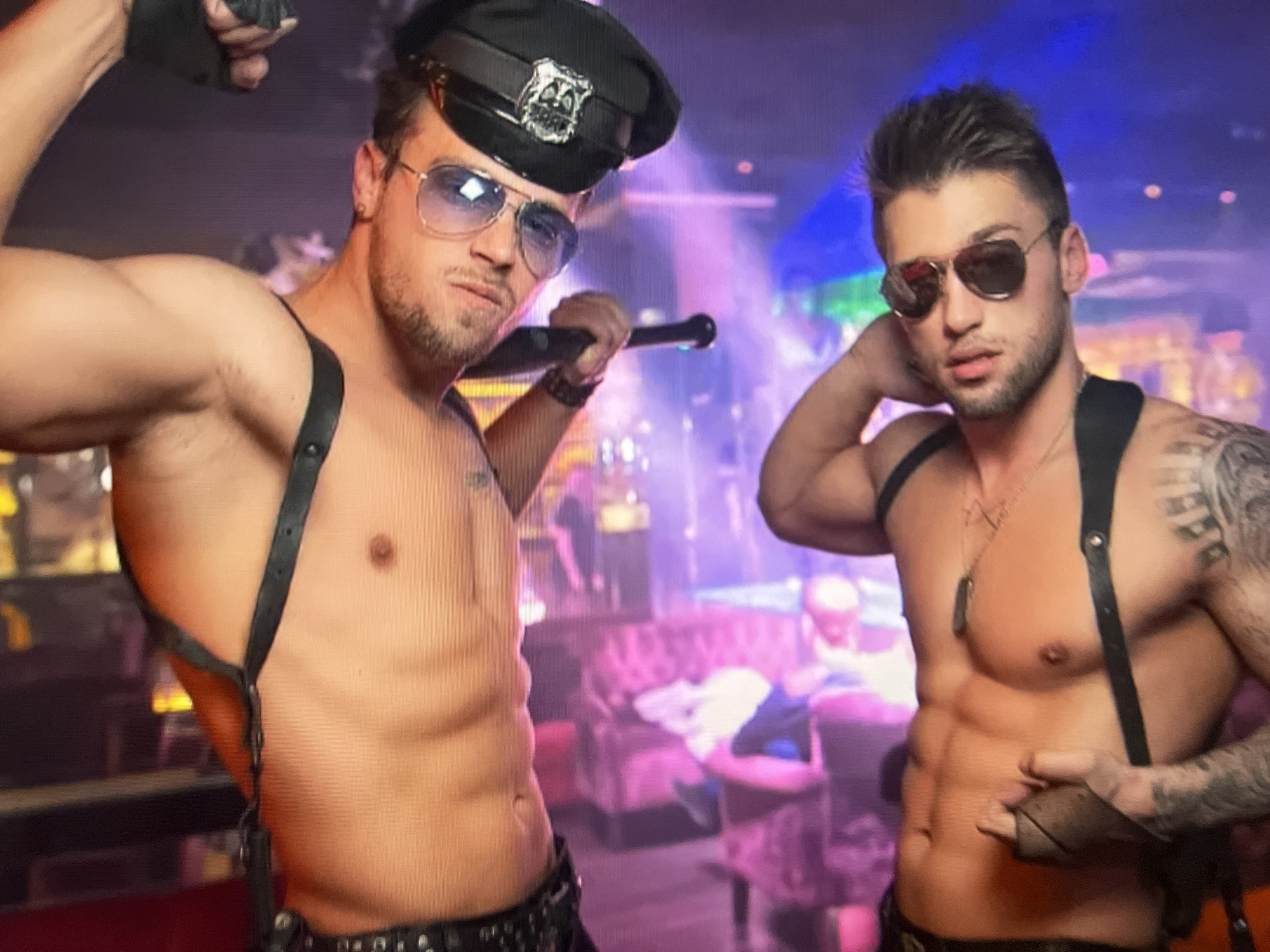 Two shirtless men, dressed in suggestive police-themed attire, are posing in a nightclub environment. The man on the left wears a police cap, aviator sunglasses, and black suspenders, holding a black baton. He is flexing his bicep. The man on the right also wears aviator sunglasses and black suspenders, with a necklace. Both men have visible abdominal muscles and are looking towards the camera. The background is dimly lit, with purple and pink lighting illuminating a lounge area with upholstered seating and blurred figures of other patrons. There is no discernible information within the image to associate it with Qarağandı city administration, Kazakhstan.