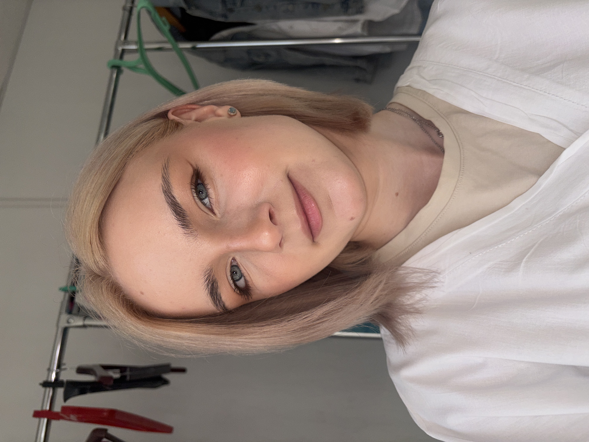 A close-up portrait shows a young woman, with the image rotated 90 degrees clockwise. She has short, light blonde or ash blonde hair styled around her face. Her eyes are blue-gray, and her eyebrows are dark and well-defined. Her fair skin shows a subtle blush on her cheeks, and her lips have a soft pink hue, suggesting natural-looking makeup.
She gazes directly at the camera with a gentle, serene expression and a slight, pleasant smile. She is wearing a light beige or tan undershirt, over which a loose-fitting white shirt or jacket is partially visible. A thin silver chain necklace adorns her neck, and a small, light-colored stud earring is visible in her right ear.
The background is softly blurred, revealing an indoor setting. A metallic clothing rack with some hanging items, including green and red hangers, can be discerned, hinting at a bedroom or dressing area. The wall behind is a light gray. The lighting is soft and even, highlighting her features. There is no visible text in the image. The context for the image is Unknown, Russia.