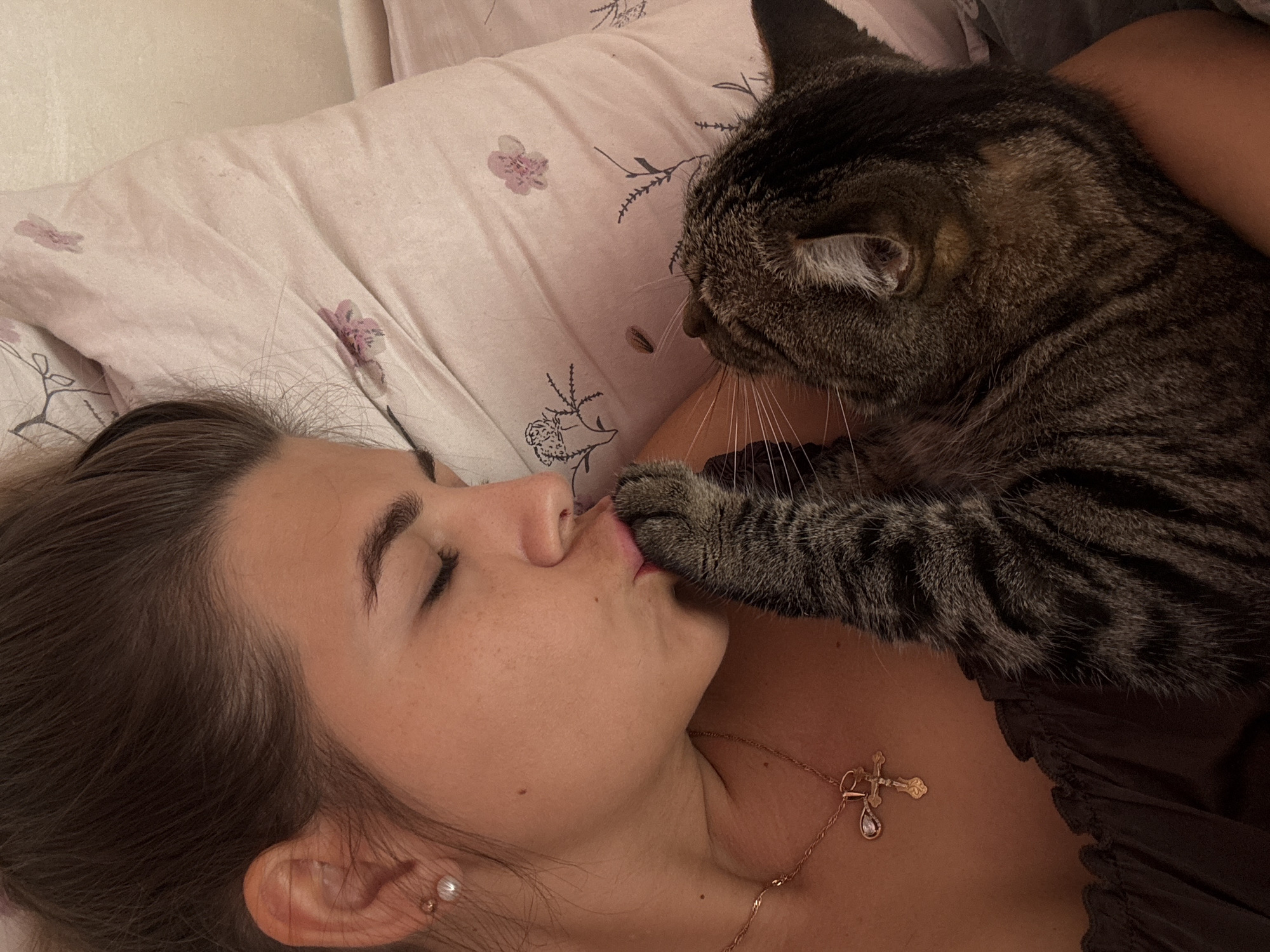 This is a close-up, eye-level indoor shot featuring a woman and a cat in bed. The woman, with her eyes closed and hair pulled back, is leaning towards the cat and appears to be kissing its paw. She is wearing a dark-colored top and a rose gold necklace with a cross pendant. The cat, a tabby, is nestled against her. The background consists of a pink pillow with a floral pattern and a plain wall. The scene appears to be taking place in a bedroom. There is no information in the image to determine its location in the city of Chaikovsky, Russia.