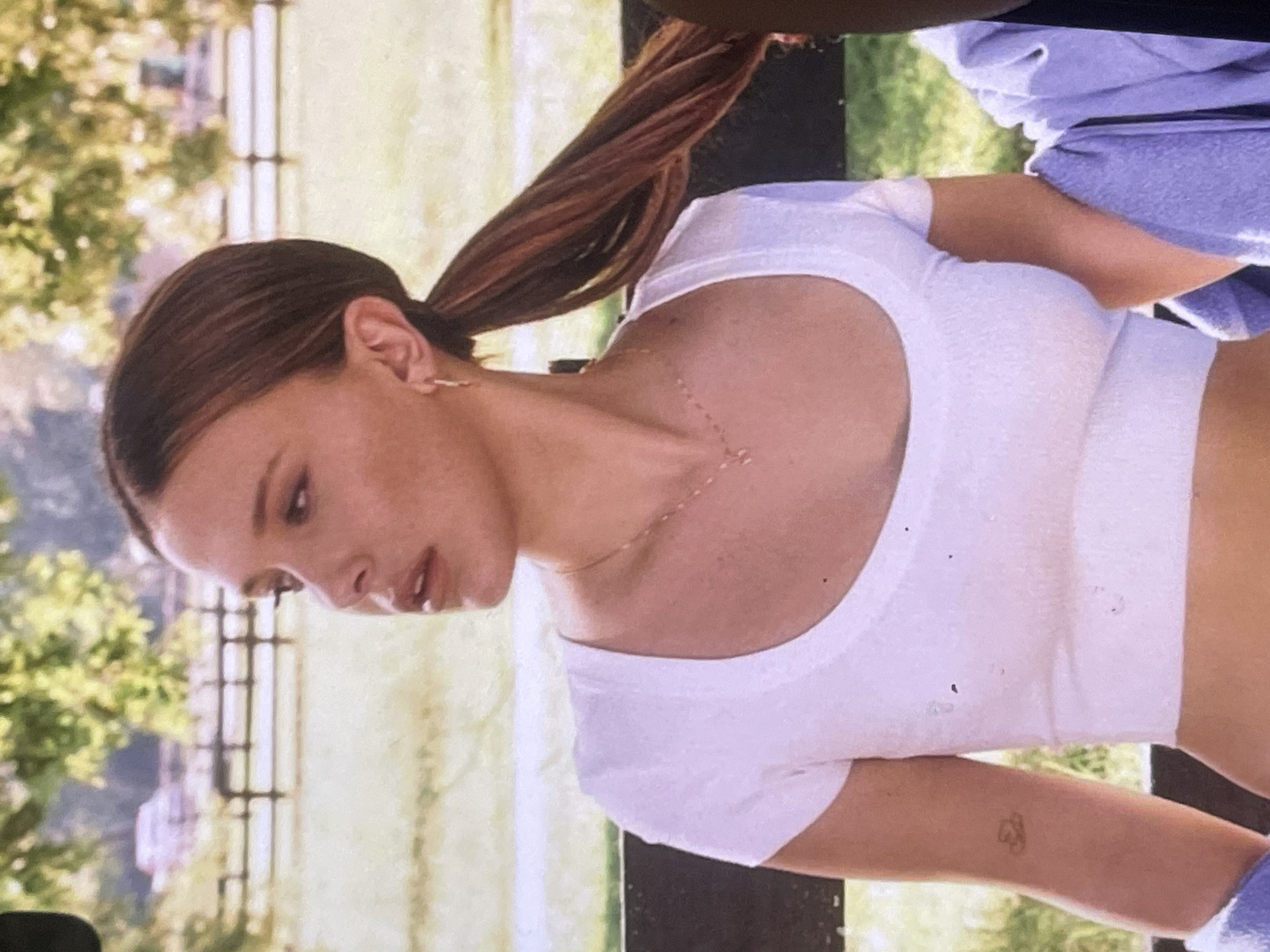 The image, initially rotated 90 degrees counter-clockwise, depicts a young woman in an outdoor setting, likely a park or garden in Poltava, Ukraine, when viewed upright. She has long, dark brown hair pulled back into a high ponytail that extends over her left shoulder. Her fair skin is dotted with numerous freckles across her face and neck. She wears a simple white, short-sleeved crop top, revealing her midriff, and a delicate gold-colored chain necklace with small, spaced elements. A small gold-colored hoop earring is visible in her left ear. Part of a lavender or light blue garment, possibly a hoodie, is draped over her right shoulder.
The woman is shown in profile, looking downwards and slightly to her left, with her lips softly parted, suggesting a contemplative or focused expression. On her left forearm, a small, faded tattoo with interconnected symbols (possibly hearts or abstract shapes) is visible.
The background is softly blurred, showing lush green foliage of trees and a light-colored fence or railings, indicating a bright daytime setting with natural light. The overall mood is serene and relaxed, with no specific activity being performed other than her standing or resting pose. There is no visible text in the image.