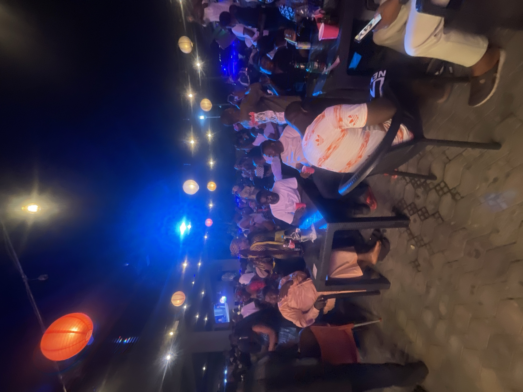 The image shows an outdoor gathering at night. People are sitting at tables, possibly dining or socializing. There are lanterns and other lights providing illumination. The location is Lokoja, Nigeria.