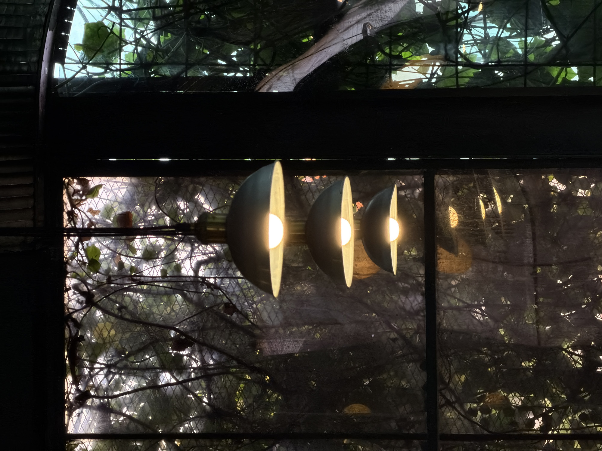 A low-angle shot through a window shows three illuminated, gray, semi-spherical lights. Behind the lights, foliage is visible through the glass, enhanced with a mesh overlay. The environment seems to be outdoors, suggested by the natural elements. The location is North Kuta, Indonesia. No people or specific actions are visible, focus is on the lighting and the background.