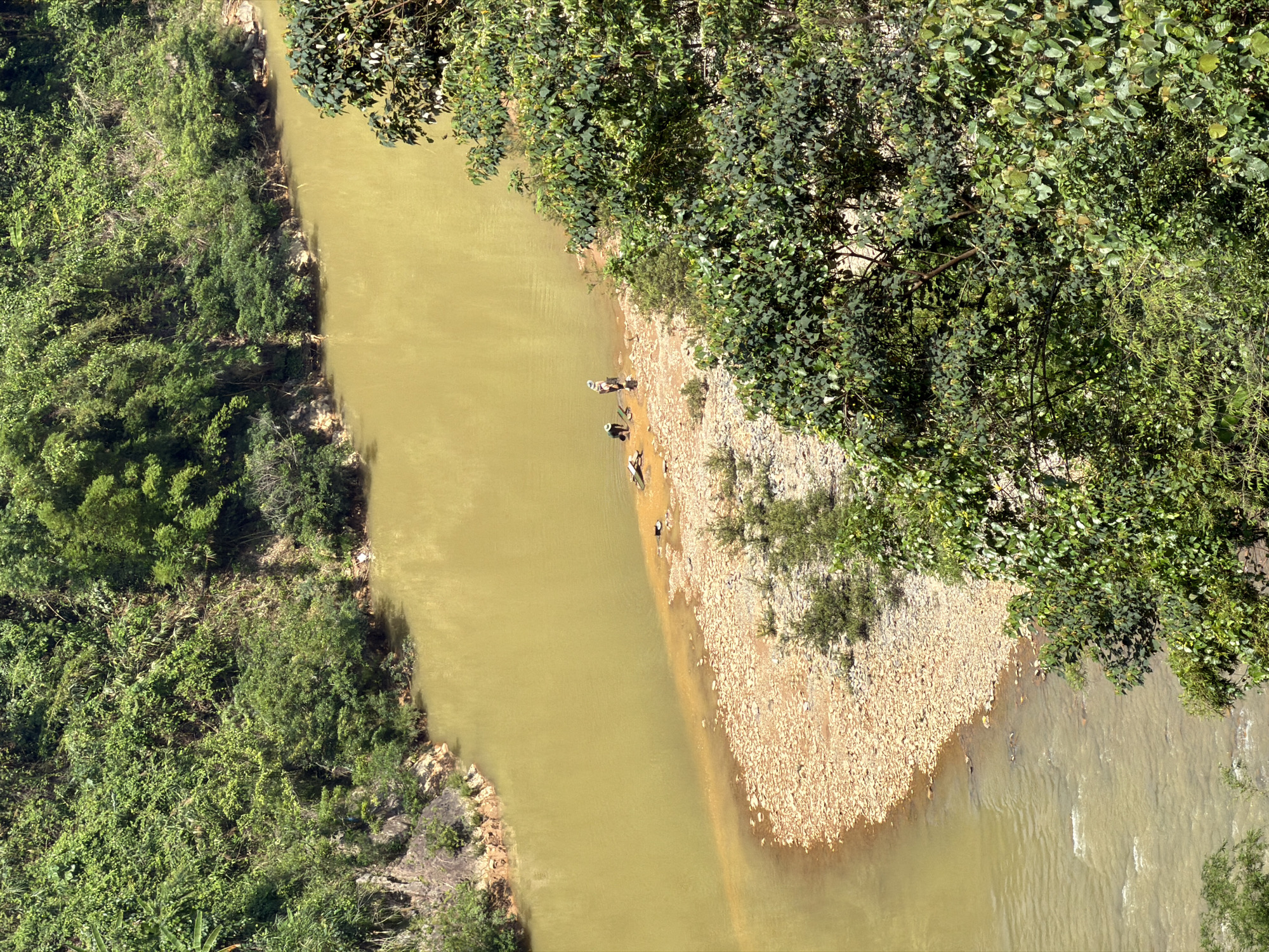 The image shows a river winding through a landscape with dense vegetation on one bank and a rocky shoreline on the other. There are several people present along the rocky shoreline, appearing to be engaged in some activity. The water in the river is murky. The location is likely Đà Nẵng, Vietnam.