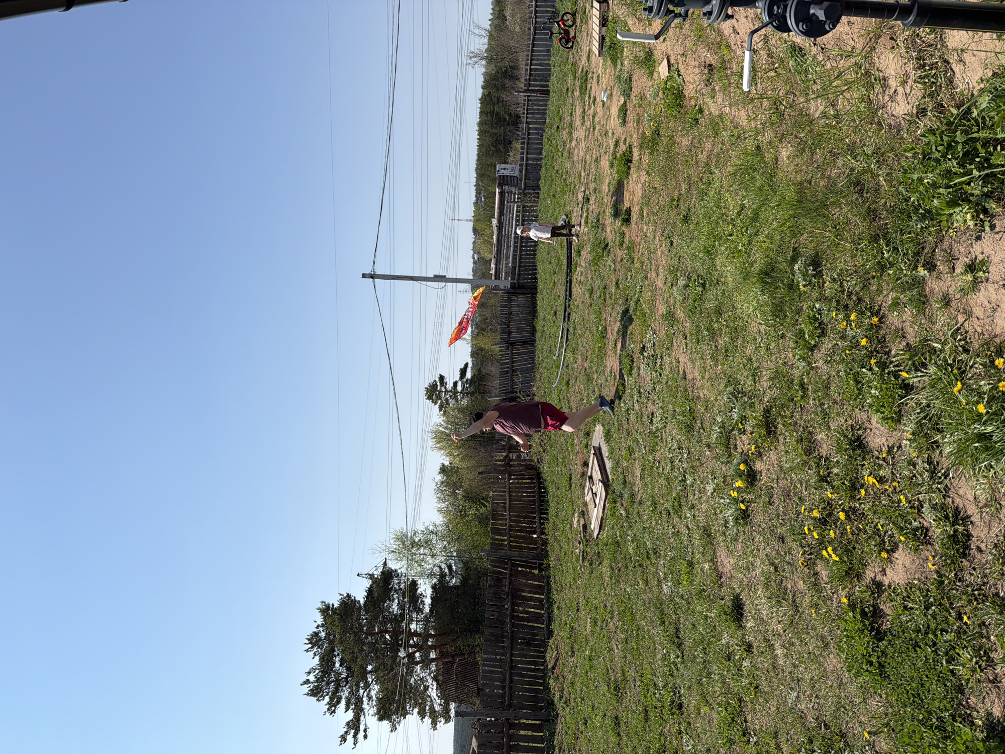 The image depicts an outdoor scene in what appears to be a yard. A person wearing a maroon shirt and shorts is standing on one leg with arms outstretched, seemingly in a balancing pose. There is a wooden fence behind them, and in the background, a child stands near an electrical pole displaying a colorful decoration. The yard is grassy with patches of bare ground and some yellow flowers. Power lines run across the sky. The location is in Chaikovsky, Russia.