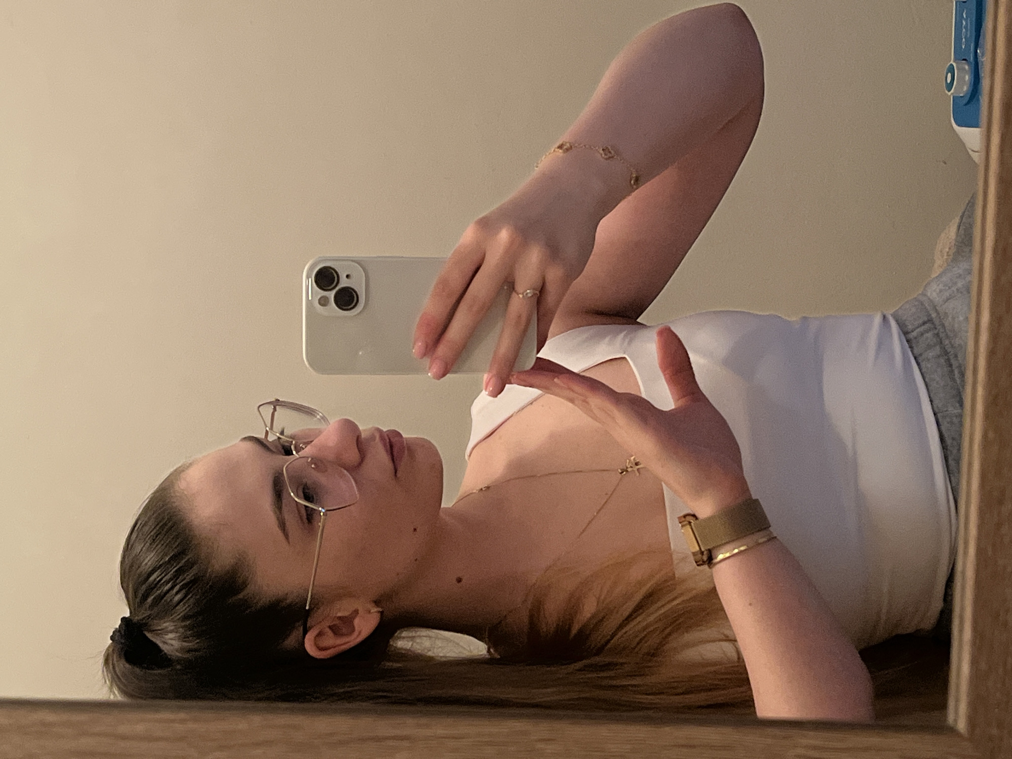 A young woman is depicted taking a mirror selfie, with the image rotated 90 degrees counter-clockwise. She has long, dark brown hair pulled back into a low ponytail or bun, and she is wearing glasses with thin, metal frames. Her expression is neutral as she looks towards the reflected camera.
She is dressed in a white sleeveless top or tank top and gray sweatpants, partially visible on her right side. Her right hand holds a white smartphone, identifiable by its triple-lens camera setup and flash, suggesting it is a modern iPhone model. A gold-colored ring is visible on her right ring finger, and her fingernails have light pink polish. Her left hand is slightly raised, with fingers splayed.
Accessories include a delicate gold-colored bracelet on her left wrist, a thin gold necklace with a small charm, and a watch on her right wrist featuring a khaki or olive green strap and a gold-toned buckle.
The background behind her is a plain, light-colored wall. A blue and white object, possibly a personal care device or container, is partially visible in the upper right corner of the frame, reflected behind her. The edges of the image show the wooden frame of the mirror. The lighting is soft and ambient, characteristic of indoor conditions. There is no visible legible text.