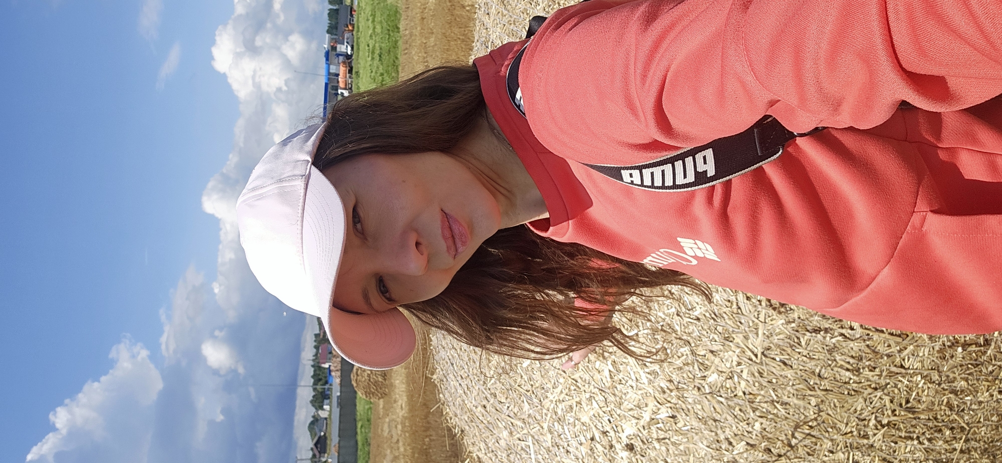 A medium shot captures a woman wearing a pink baseball cap and a coral-colored sweatshirt, taken from a slightly low angle. She is positioned in front of a large hay bale, which takes up a significant portion of the foreground. The background displays a rural landscape under a bright sky with scattered clouds, featuring a field with more hay bales, some buildings, and vehicles in the distance. The scene appears to be set in a rural area, possibly near agricultural fields, which could be located in or around Лятчанскі сельскі Савет, Belarus, given the context. The woman is looking towards the camera with a slight smile, and her right arm is extended forward.