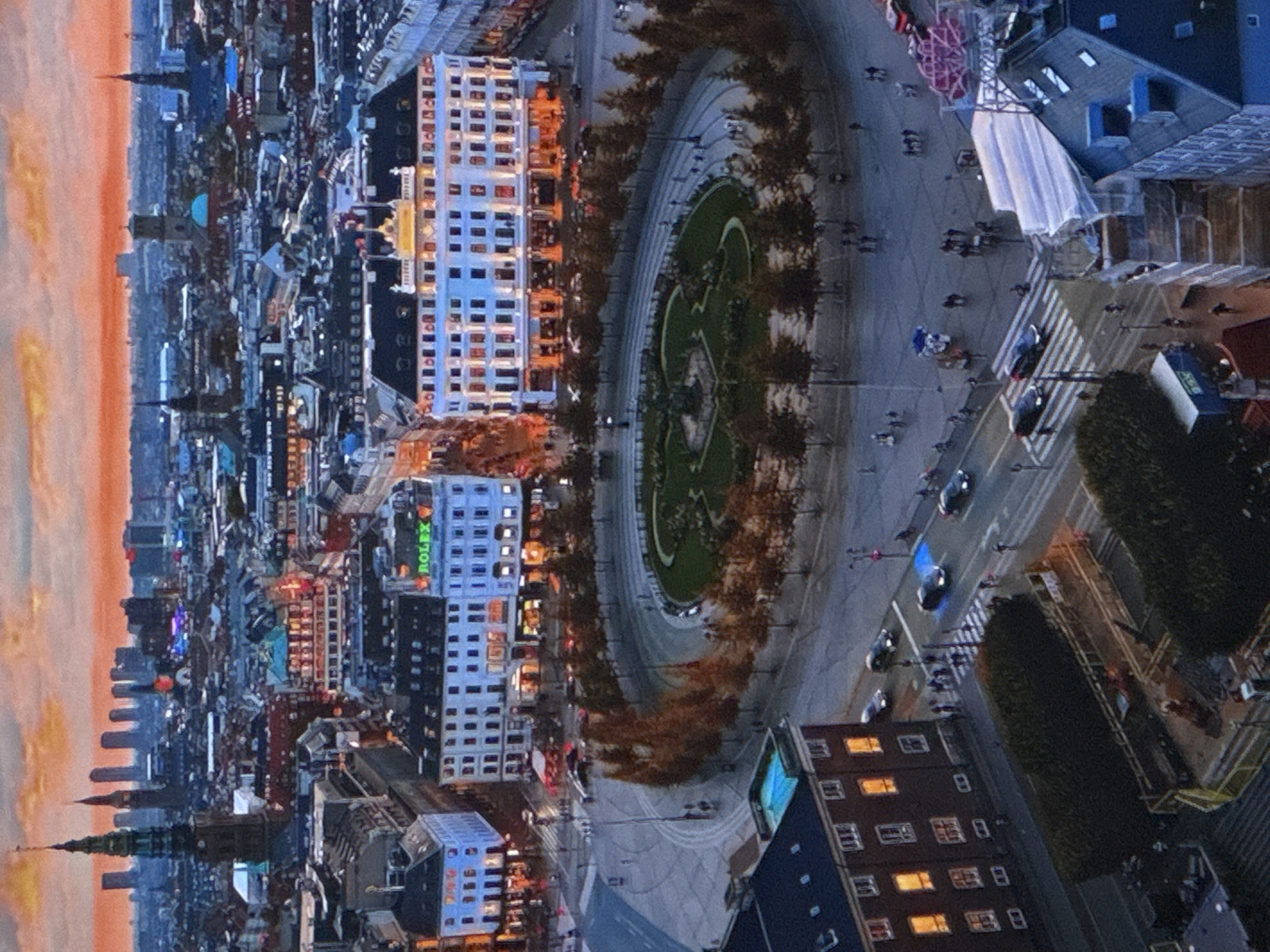 This is an aerial view of a city square at dusk. In the background, a skyline of tall buildings is visible against an orange and pink sky. The foreground features a circular park with trees and a monument at its center. Surrounding the park are several large, ornate buildings, some with illuminated windows. A street with cars and pedestrians curves around the square. One building displays a prominent "ROLEX" sign. Another building has a rooftop swimming pool. Construction is visible on the lower right side of the image. The overall scene depicts a bustling urban environment transitioning into evening.