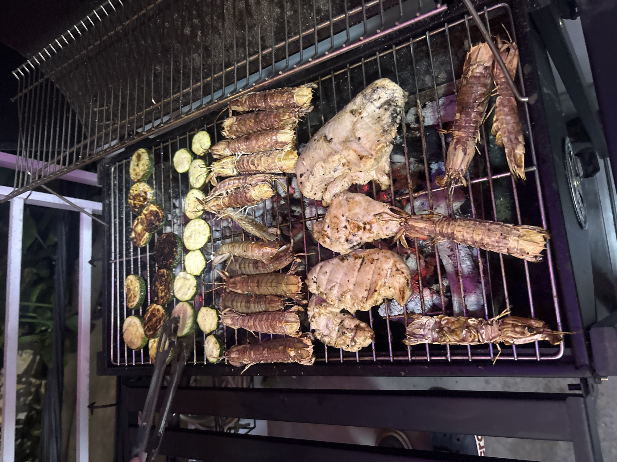 The image shows a barbecue grill with food cooking on it. The food consists of mantis shrimp, grilled chicken, and sliced zucchini. The grill is located outdoors, possibly in Nha Trang Ward, Vietnam. Tongs are present on the grill, presumably being used to move the zucchini.