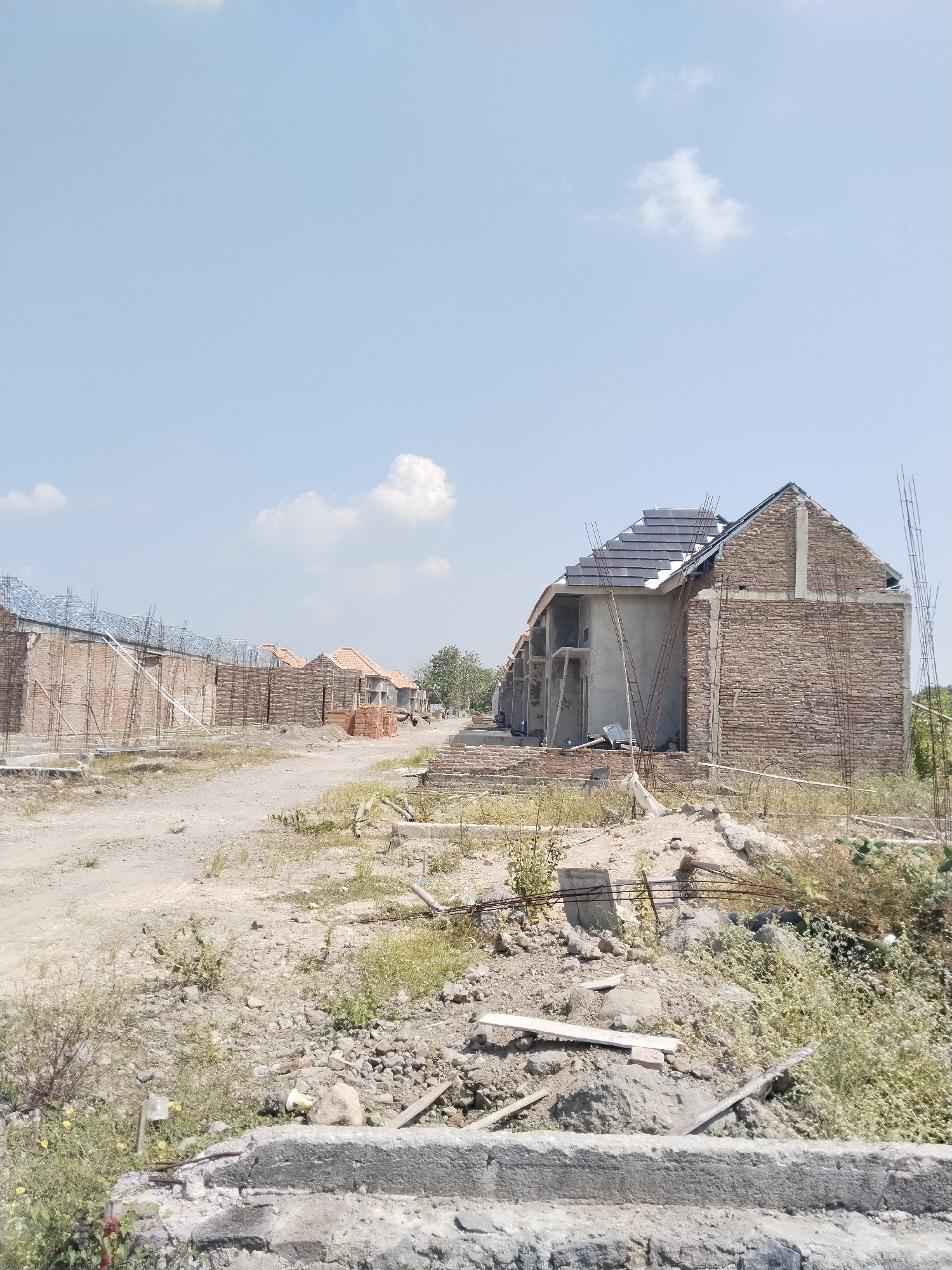 The image displays an outdoor scene of a residential development under construction, likely taken on a bright, sunny day. A wide, unpaved dirt road stretches from the foreground into the background, flanked by houses in various stages of completion.
In the immediate foreground on the right, a house exhibits exposed brick walls and a partially installed roof with dark gray tiles. Concrete structural elements and rebar protrude from the unfinished structure. The ground around it is rough, rocky, and covered with sparse, dry weeds and some construction debris, including concrete fragments, wooden planks, and plastic waste.
Further down the dirt road on the left, a row of similar houses is visible. Many of these structures show only their basic brickwork and rebar frameworks, while others have begun to receive red or orange roof tiles. Piles of bricks are stacked near some of these buildings, indicating active construction. The landscape on both sides of the road consists of unkempt, dry patches of grass and dirt.
The sky above is a clear, bright blue with a few scattered white clouds, suggesting fine weather and ample daylight, possibly midday. No people are visible anywhere in the scene, and there is no discernible text. The overall setting is that of a developing residential area, characterized by new construction and an unfinished, raw environment.