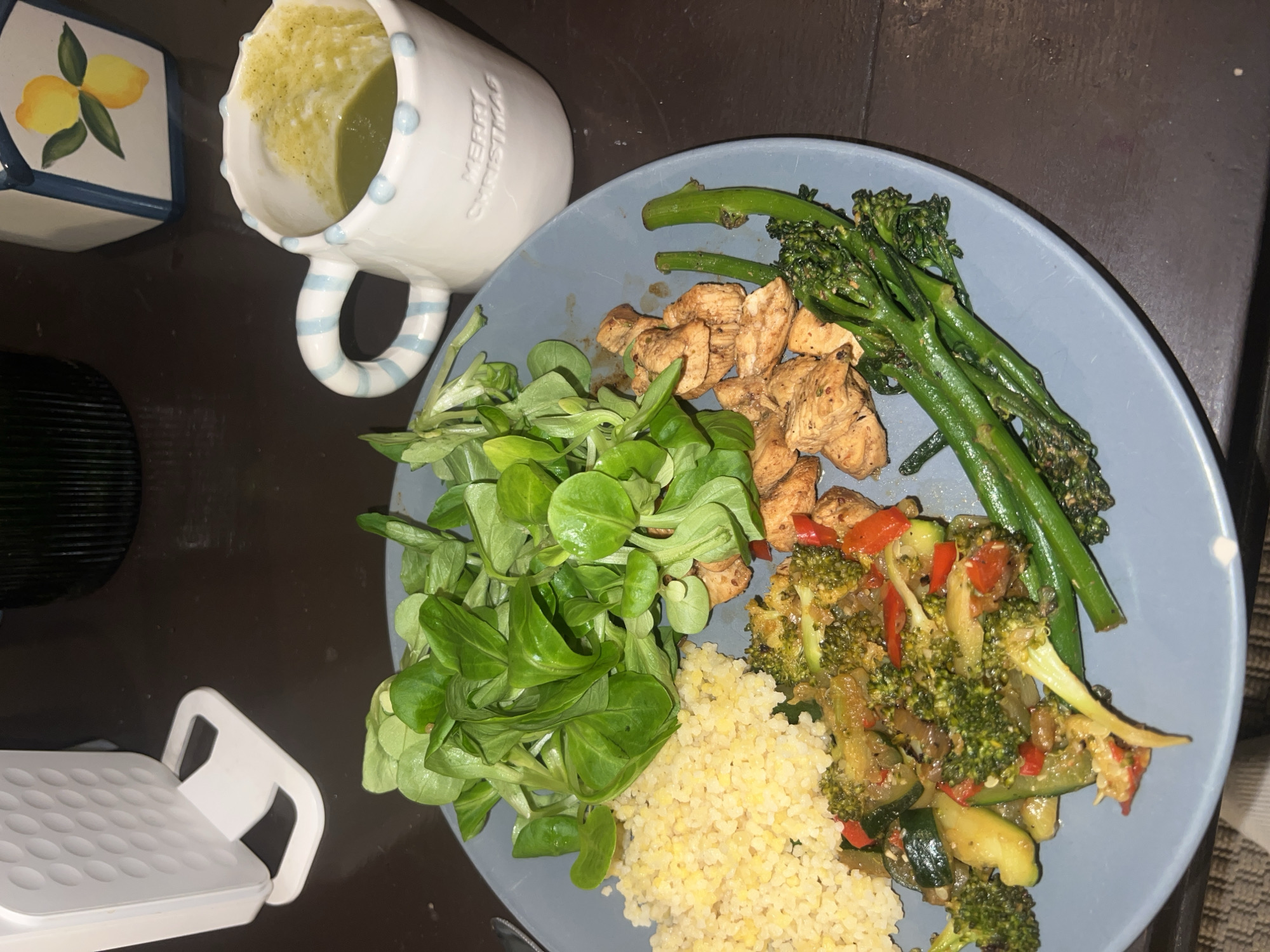 The image depicts a plate of food and a mug, placed on a dark surface. The plate contains mâche lettuce, chicken pieces, cooked bulgur wheat, sautéed vegetables (broccoli, zucchini, red bell pepper), and cooked broccoli. The mug appears to hold a green liquid, possibly soup. To the left of the plate is a small, square ceramic container decorated with lemons. A white object, possibly a kitchen appliance, is visible in the lower left corner. The setting appears to be a dining area, but it is not possible to identify any particular location in Paris from the information in the image.
