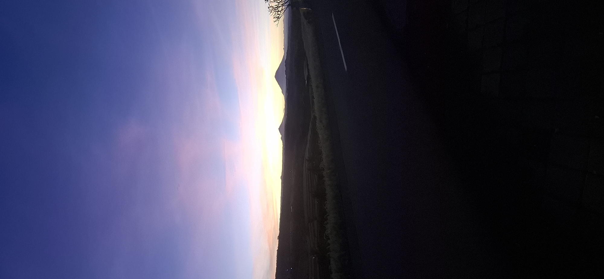 The image shows a landscape in Penebel, Indonesia, during the late afternoon or early evening. The sky is a gradient of deep blue to pale pink and yellow near the horizon, suggesting either sunrise or sunset. A mountain range is visible in silhouette against the bright horizon. A road with a visible white line runs alongside agricultural fields. The foreground is dark, possibly indicating a building or dense foliage, obscuring details.