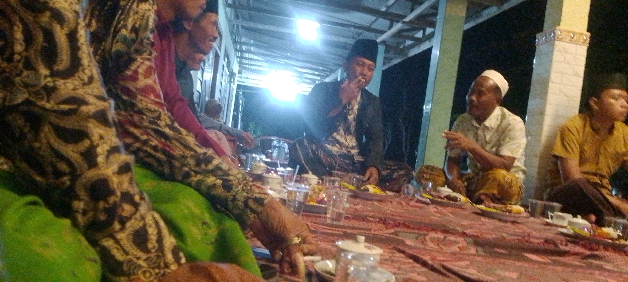A group of men are gathered around a low table, possibly for a meal or a social gathering, at night. The setting appears to be outdoors or in a semi-enclosed space with a corrugated roof and open sides, illuminated by bright artificial lighting. The men are dressed in traditional attire, including sarongs and head coverings. One man in the center is wearing a black cap and jacket and is smoking. Another man on the right is wearing a white patterned shirt and a white skullcap. A third man on the far right is wearing a yellow shirt. The table is covered with a patterned cloth and laden with plates of food, cups, and glasses. The overall atmosphere suggests a casual and communal event.