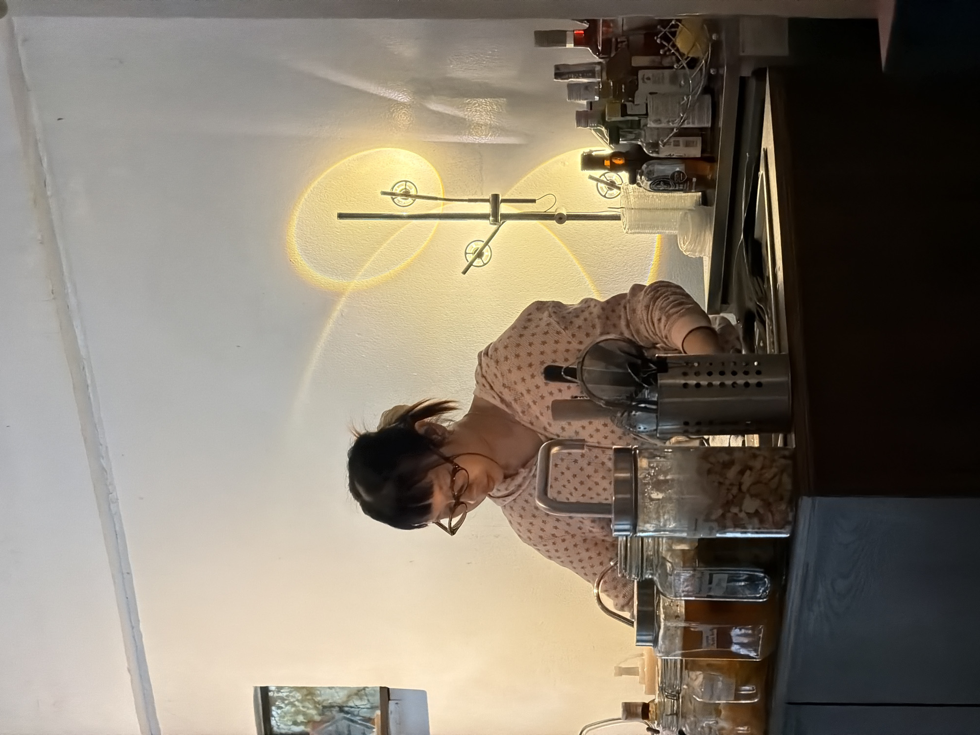 This image, taken in Chiang Mai City Municipality, Thailand, shows a woman with dark hair, glasses, and a pink patterned shirt, leaning over a kitchen counter. She is engaged in an activity, possibly preparing food or organizing items, as her hands are near several glass jars filled with ingredients and a stainless steel utensil holder.
The setting appears to be an indoor kitchen area, with a white, textured wall and a ceiling illuminated by soft, warm light. Two circular light fixtures on the wall cast distinctive, swirling patterns of light. A shelf above the counter is stocked with various bottles, suggesting a well-equipped space. To the right of the woman, a dark wooden cabinet or countertop occupies the frame, with another dark surface below it.
The lighting suggests it might be evening or a dimly lit interior. The overall mood is calm and focused, with the woman engrossed in her task. A small framed image is visible in the lower left corner, partially obscured by shadow, offering a glimpse of an outdoor scene. There is no visible text in the image.