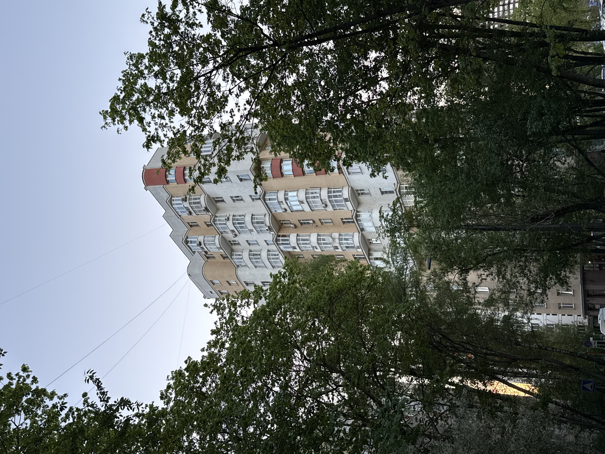 The image shows a tall residential building in Moscow, Russia, partially obscured by trees. The building has a unique architectural design with rounded balconies and alternating color bands on the facade. The sky is a pale blue, suggesting early morning or late evening. Power lines stretch from the top of the building. Trees are in full leaf, indicating it is likely spring or summer. Other buildings and street signs can be seen in the background, further suggesting an urban environment.