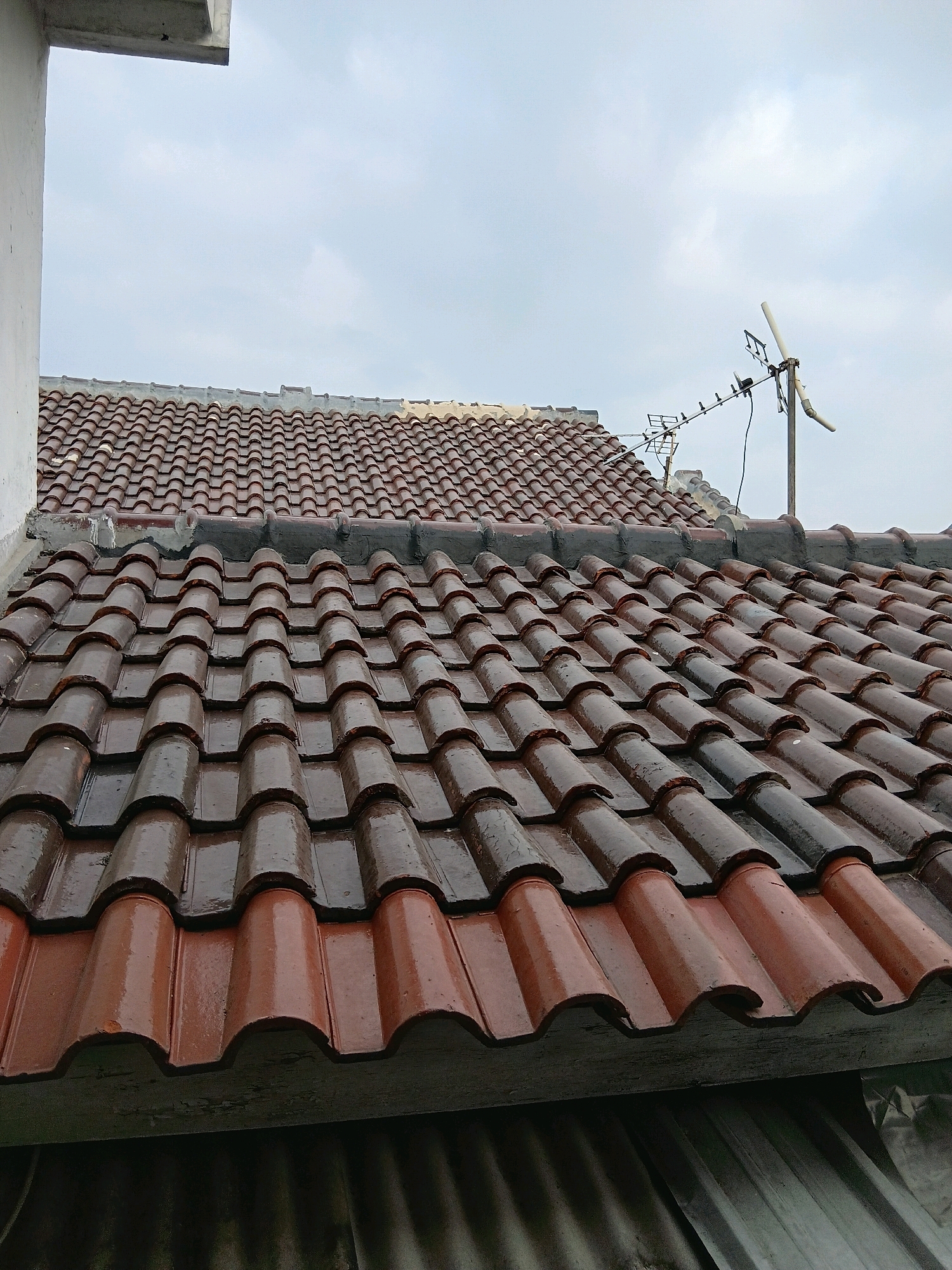An overcast sky looms above a close-up, high-angle shot of a tiled rooftop. The tiles are a mixture of brown and reddish-brown, and appear wet, reflecting the dull light. They are laid in a wavy pattern, typical of many Indonesian rooftops. To the right of the image, a television antenna is mounted on a pole, its metal structure stark against the cloudy sky. In the background, another tiled roof is visible, similar in style. To the left of the frame, a portion of a white wall extends upwards, its clean surface contrasting with the textured tiles. The overall scene suggests a residential area in Cirebon, Indonesia, under a damp, cloudy day.