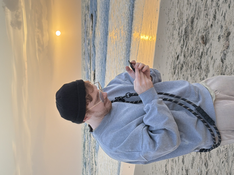 A solitary individual stands on a sandy beach in Tel-Aviv, Israel, during sunset. The person, appearing male with reddish-brown hair and light stubble under a black knit beanie, wears clear-framed eyeglasses, an oversized light grey crewneck sweatshirt, and light beige corduroy pants. A black and white striped rope strap with black plastic elements is slung diagonally across their torso. Both hands hold a black smartphone, with fingers positioned as if interacting with the screen. The sun, a bright orange disc, is low over the western sea horizon, casting a prominent reflection on the moderate waves. The sky is a gradient of orange and yellow with scattered clouds. The beach features light-colored sand, with a small dark object, potentially organic matter, visible on the sand in the lower right quadrant.