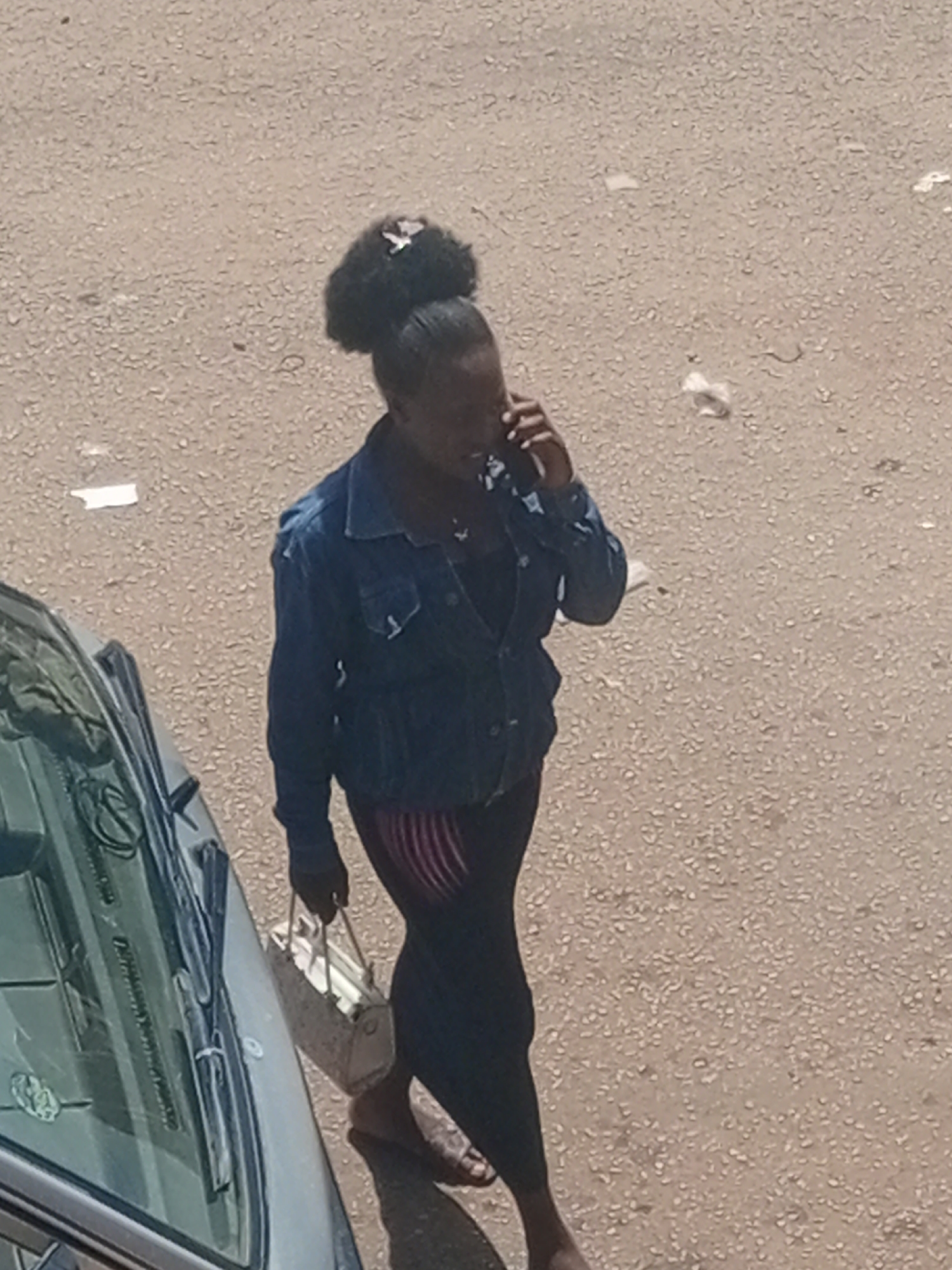 In Bukuru, Nigeria, a woman with dark skin and her hair styled in a high bun with a white accessory walks on a paved surface, possibly a street or sidewalk. She is wearing a blue denim jacket over a dark top and dark pants with a red and white striped pattern on the lower legs. Her right hand is holding a light-colored handbag, and her left hand is raised to her ear, as if speaking on a mobile phone. She is wearing sandals. To the left of the woman, a portion of a dark-colored car is visible, showing part of its windshield and hood. The ground is a textured, light brown or beige surface with small pebbles and some scattered white debris. The lighting suggests it is daytime, with the sun casting shadows.
