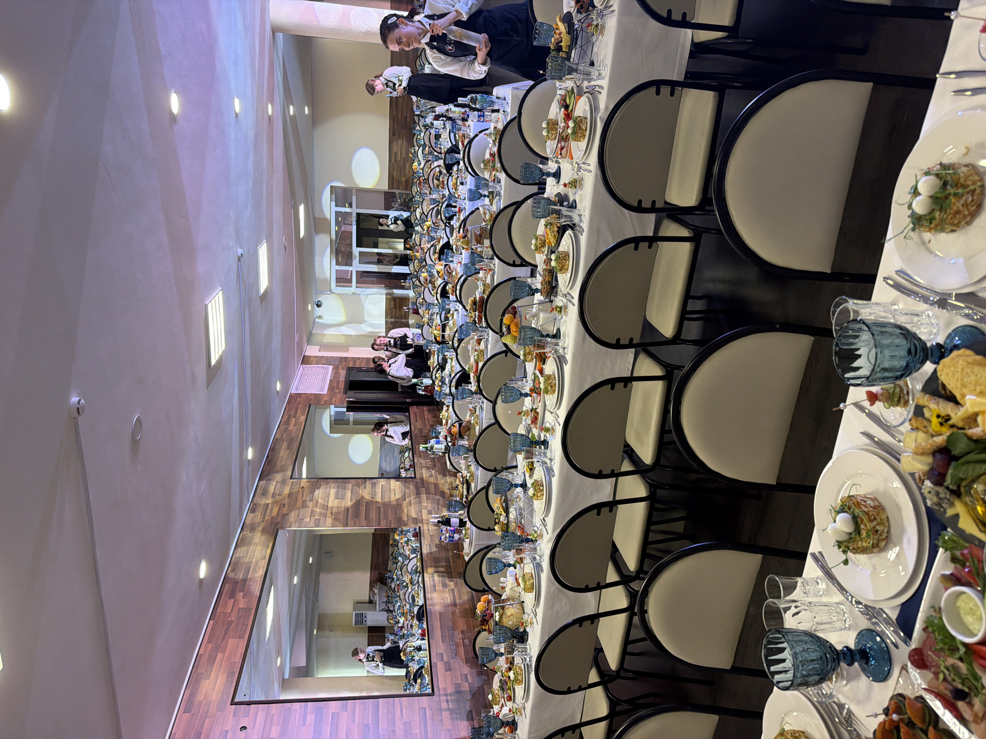 The image depicts a long, elegantly set banquet table, likely in a restaurant or event hall in Chaikovsky, Russia. The table is adorned with white tablecloths, numerous plates of food, silverware, and blue glassware. Several servers, dressed in black aprons and white shirts, are attending to the table and interacting with guests who are not clearly visible. The setting appears to be indoors, with warm lighting and a polished wooden floor. The overall atmosphere suggests a formal event or celebration, possibly a wedding reception or a catered dinner, given the extensive setup and the presence of multiple servers. The lighting is artificial, indicating it could be evening or a dimly lit venue. Mirrors on the wall reflect the scene, creating a sense of depth and mirroring the organized arrangement of the dining setup. The mood appears professional and organized, with the servers focused on their tasks. There is no visible text in the image.