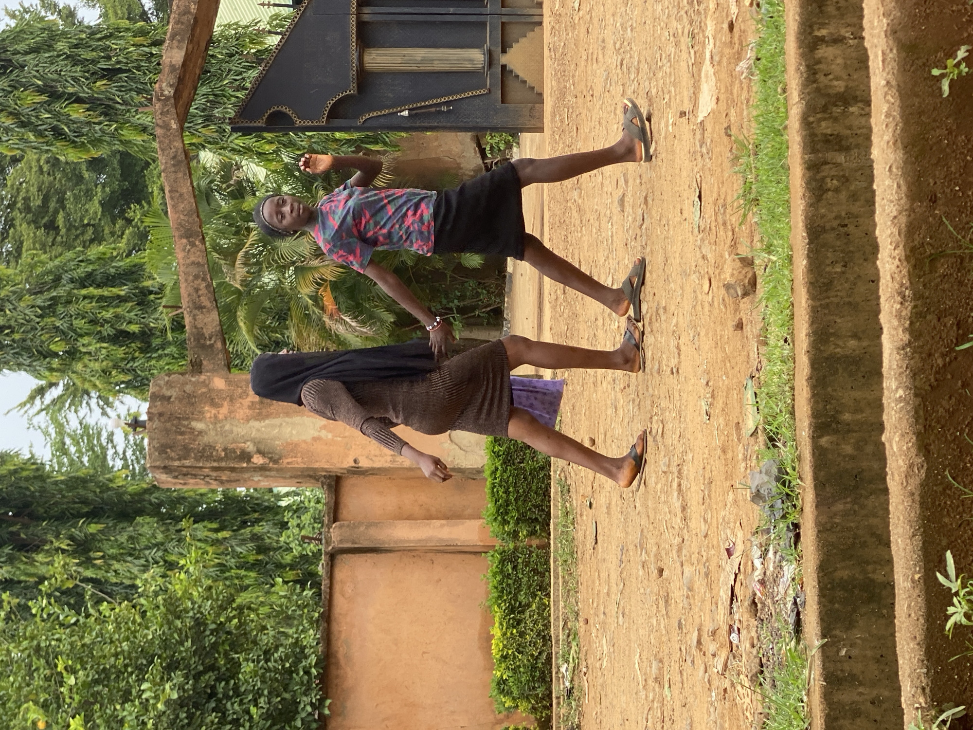 Two individuals, a boy and a girl, are standing on a dirt path. The boy is wearing a patterned shirt and black shorts. The girl is wearing a brown dress, with a black head covering. They are holding hands. The setting appears to be outdoors with a gate, trees, and a building in the background. Trash is visible along the edge of the path. It is daytime. The location is speculated to be Maigizo, Nigeria.