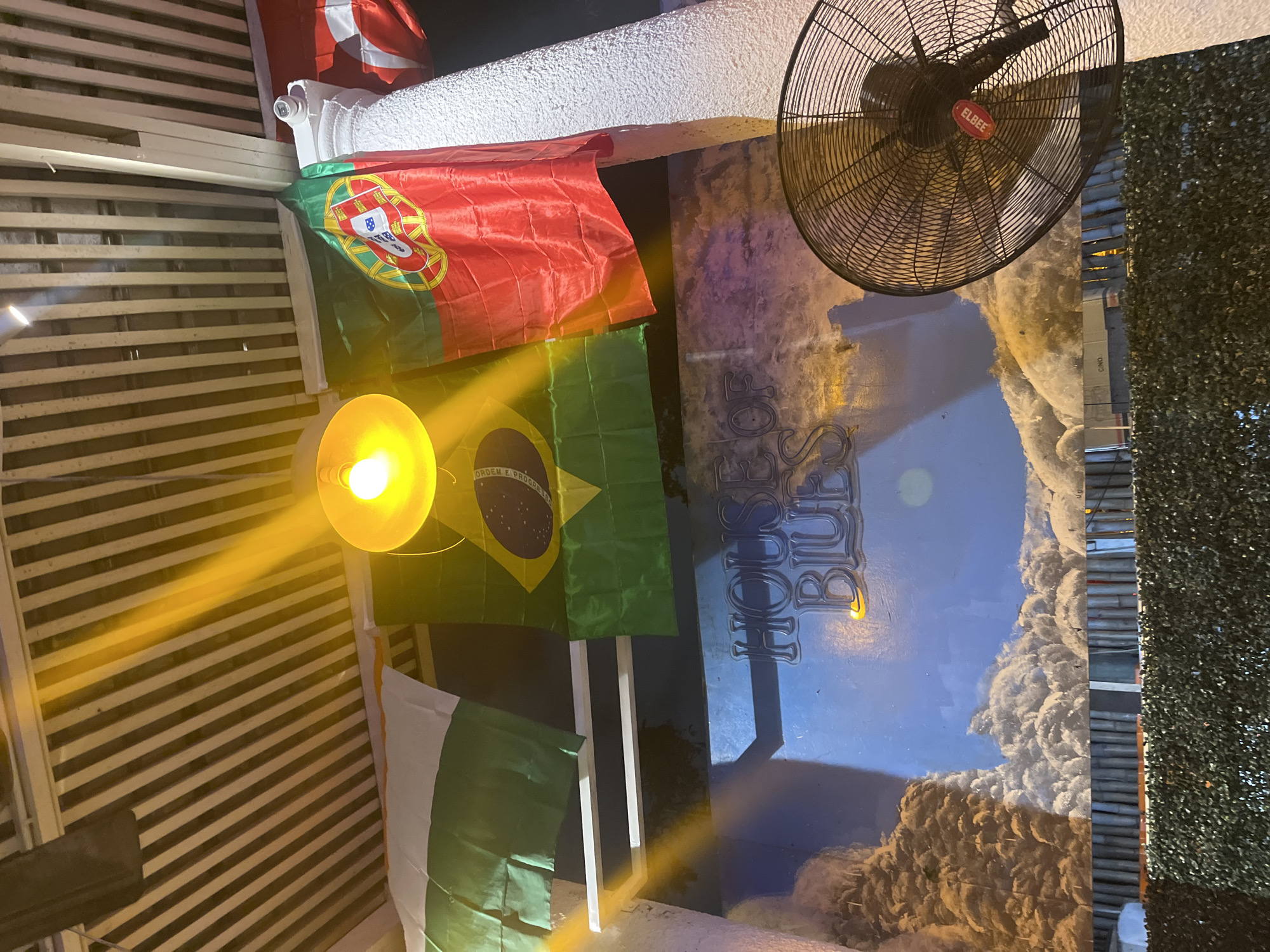 The image captures an outdoor scene at the "House of Blues" venue in Accra, Ghana. Flags of India, Brazil, and Portugal are displayed, suggesting an international theme or celebration. A bright yellow lamp hangs from the slatted ceiling, casting a warm glow. Below the flags, a blue mural depicts a sky with clouds. A large black fan with the "ELBEE" logo is positioned on the right side of the frame. The overall atmosphere appears festive and possibly related to a sporting event or cultural gathering.