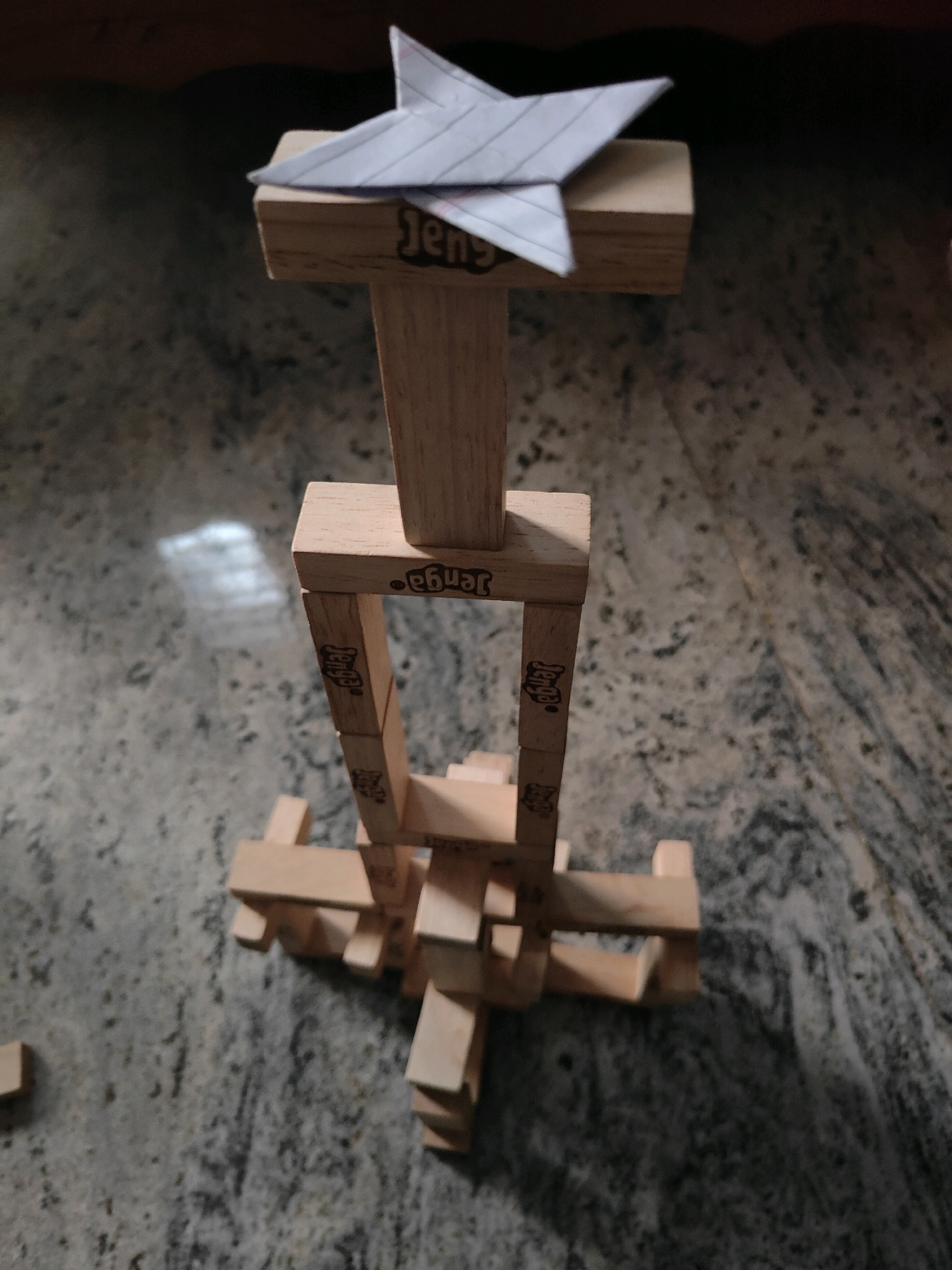 An origami star, made from lined paper and folded into a traditional pentagonal shape, rests atop a Jenga tower. The Jenga tower, constructed from approximately 25 wooden blocks, is built on a grey, speckled countertop. The tower has a central vertical section, topped by two blocks oriented perpendicularly, and a wider base made of horizontal blocks. The origami star is positioned directly on top of the uppermost Jenga block. The image is taken from a high-angle perspective, looking down onto the tower and star. The background is blurred, showing a wooden surface and suggesting an indoor setting. There are no people or animals present in the image. The lighting appears to be artificial, with a soft glow illuminating the scene. There is no specific indication of the city of Karapalli, India, within the image itself.