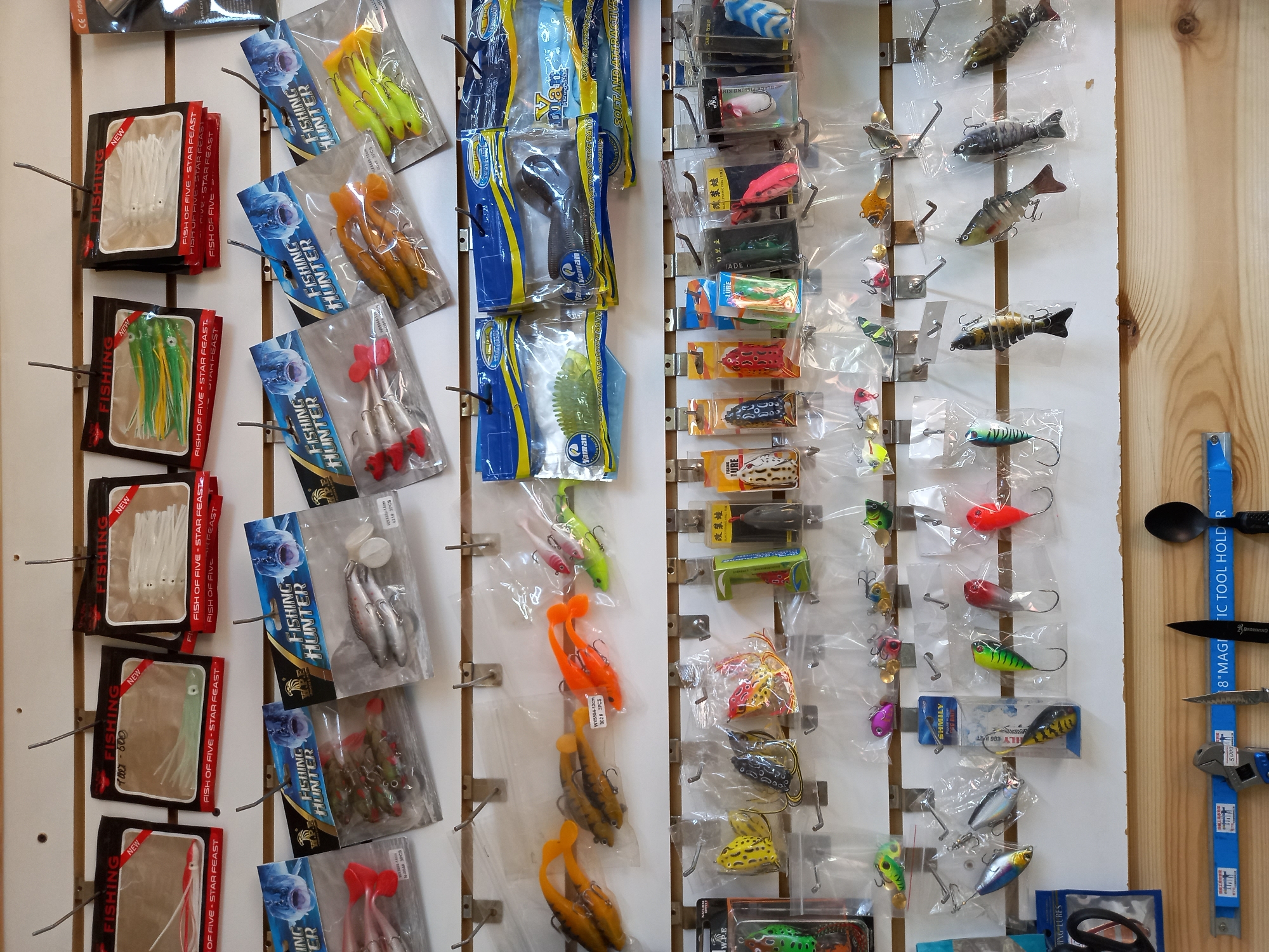 The image shows a pegboard display of fishing lures. The lures are packaged in plastic and hung on metal hooks. They vary in type, color, and size. Brands visible include "Fishing Hunter" and "Fish of Five." A wooden strip to the right displays a magnetic tool holder with a spoon, knife, and pliers.