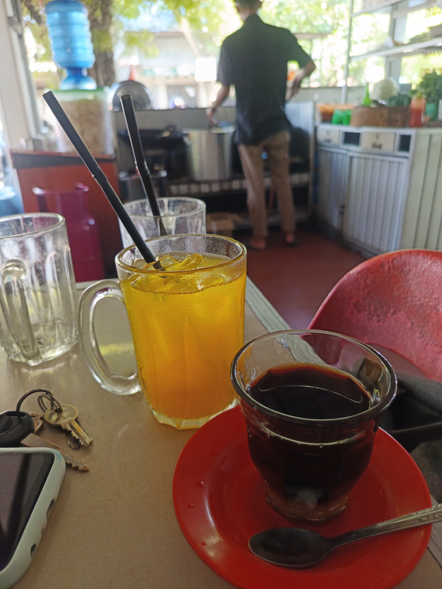 This image captures an interior scene of an informal eatery or cafe in Lhokseumawe, Indonesia, illuminated by natural daylight.
In the immediate foreground, a light brown tabletop displays several objects. To the left, a white smartphone with a light blue-green protective case rests beside a set of keys, including a gold-colored key and one attached to a black "HONDA" keychain. Moving right, an empty clear glass mug with vertical ridges is present. Adjacent to it is a larger clear glass mug with a handle, filled with a bright orange-yellow liquid containing ice cubes and a black drinking straw; condensation is visible on its surface. Behind this drink, a partially obscured third clear glass also contains a black straw. Further to the right, a smaller clear glass holds a dark brown-black liquid, consistent with coffee, resting on a red circular saucer. A small, metallic spoon is placed on the saucer to the right of the glass. The upper portion of a red plastic chair back is visible at the far right.
In the midground, a person stands with their back to the viewer, positioned behind a counter in the center-left of the frame. They are wearing a dark, short-sleeved shirt and light brown trousers, with their arms slightly raised, indicating an ongoing activity at the counter. The counter features a light-colored, possibly metallic, slatted front and holds various items on its surface, including a light brown woven basket, a green container, and a red squeeze bottle. The floor in this area is a reddish-brown hue.
The background is brightly lit, revealing green foliage outside, suggesting an open window or an outdoor seating area. A large, blue upright water dispenser is partially visible in the far-left background. The overall environment appears casual and functional, bathed in natural light.