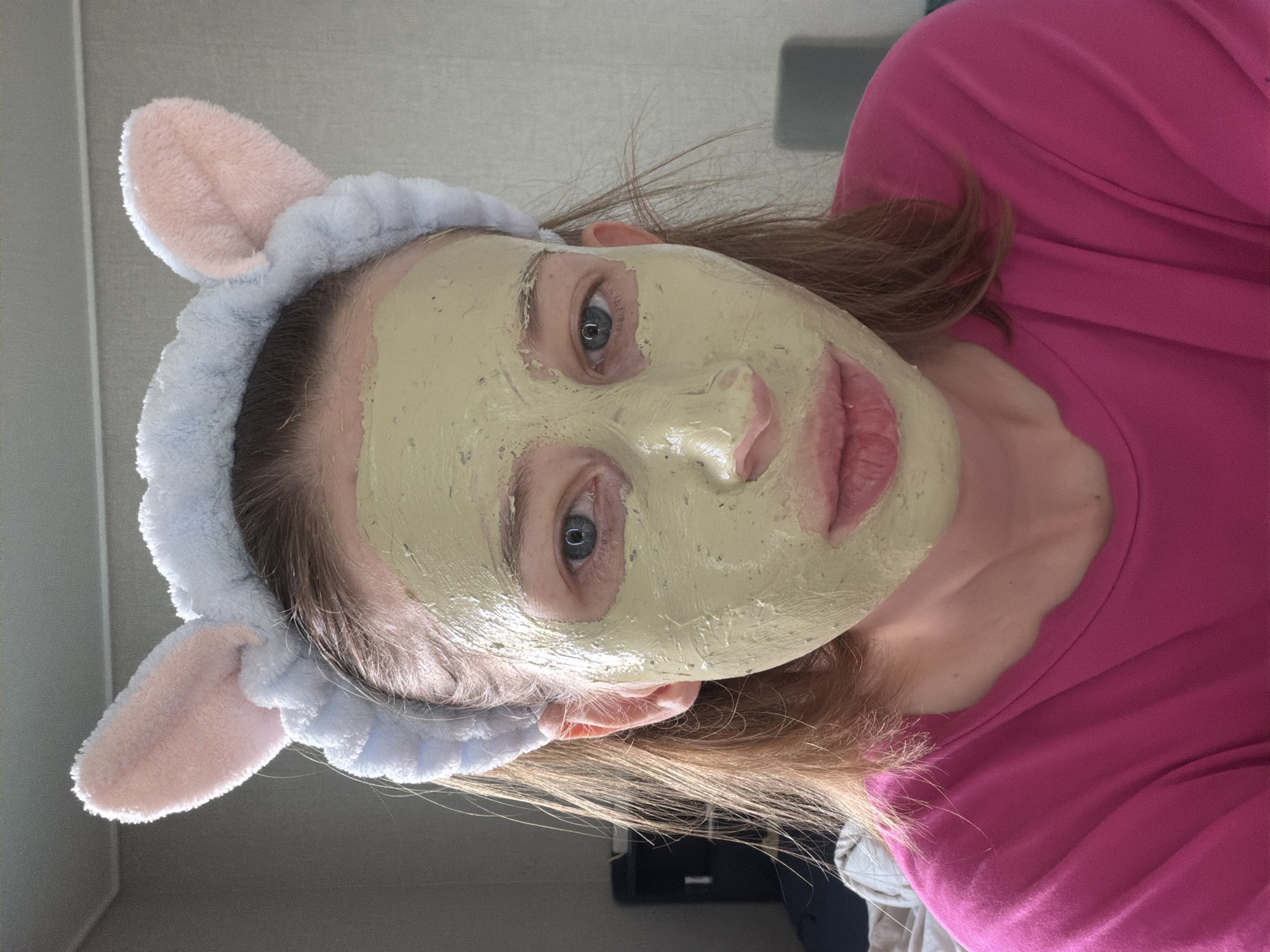 A young woman is shown in a close-up portrait, wearing a light blue headband with pink bunny ears. Her face is covered in a light green clay mask. She has light brown hair that falls around her shoulders. She is wearing a bright pink t-shirt. The background appears to be an indoor setting with a plain, light-colored wall. The image is a selfie, suggesting the woman is likely at home or in a personal space. The setting is consistent with an urban environment, potentially within Dongducheon-si, South Korea, given the user prompt.