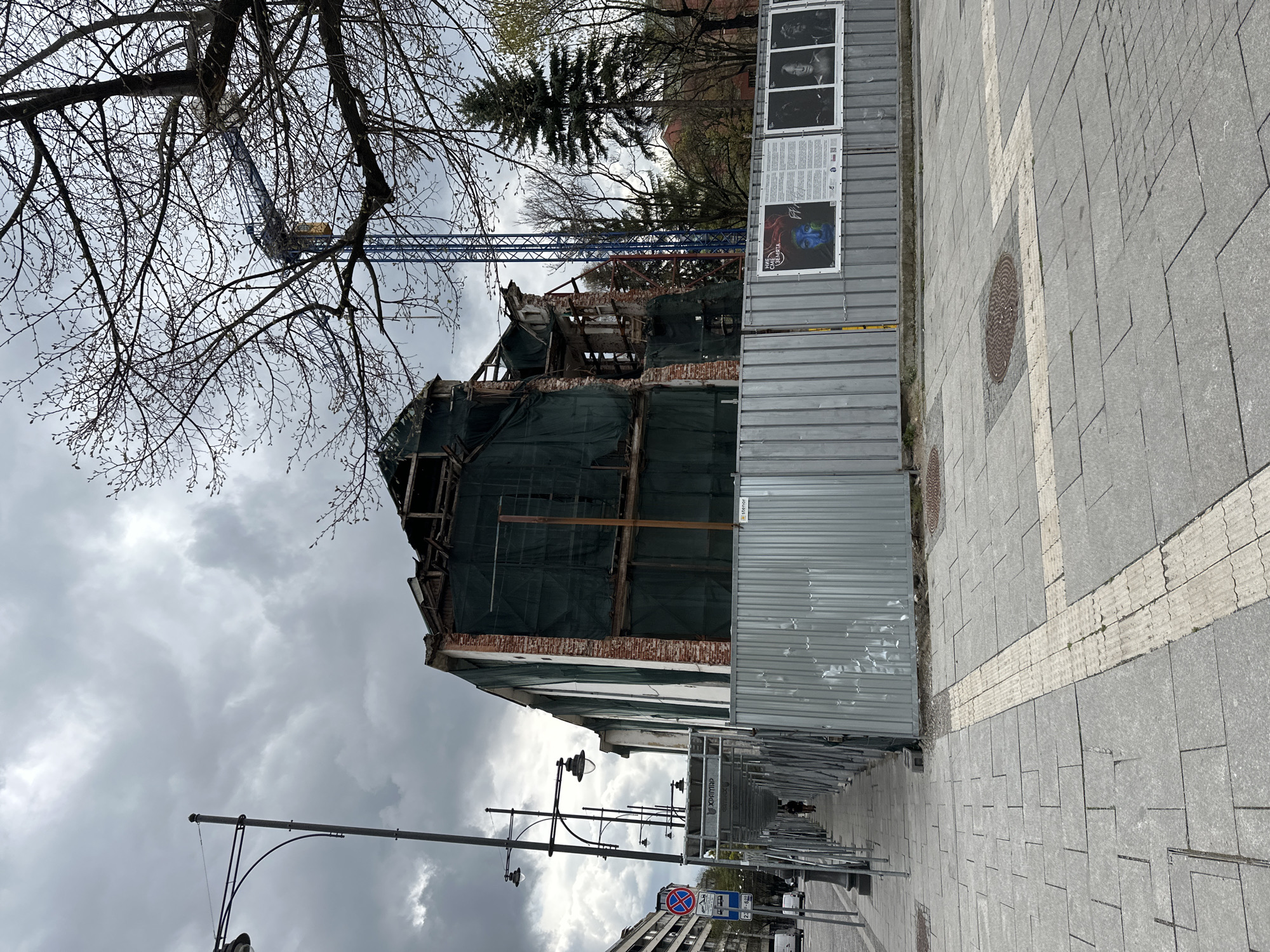 The image presents an urban scene in Sofia, Bulgaria, depicting a building under construction or demolition, flanked by a public sidewalk and street. The building's upper floors are significantly dismantled, showing exposed wooden beams and brickwork, partially covered by dark green construction netting. A tall blue construction crane is visible behind and above the structure, with its boom extending into the cloudy sky.
A corrugated metal fence, acting as a temporary barrier for the construction site, runs along the sidewalk. Several posters are attached to this fence. One poster displays a black and white portrait with the name "Phoebe" underneath, while another features an abstract design of a blue and red face with the text "WE CAN CHANGE THE WORLD" and a globe-like symbol. Further along, a small white sign on the fence reads "Stercor".
The sidewalk is paved with grey square tiles, incorporating a lighter-colored tactile strip for the visually impaired. Manhole covers are embedded in the pavement. Beyond the main construction fence, a covered walkway made of scaffolding, bearing the Cyrillic text "ХХАРЯМР" (likely a company name like "Kharyamar"), extends down the street, providing shelter for pedestrians. A few distant figures can be seen walking under this structure.
The weather appears overcast, with thick grey clouds dominating the sky. Bare tree branches, possibly with emerging buds, frame the upper part of the scene. Streetlights with decorative fixtures are positioned along the sidewalk. A blue circular traffic sign with a red diagonal line, indicating "No stopping" or "No parking," is visible further down the street. The scene suggests daytime during late winter or early spring, characterized by ongoing urban development.
