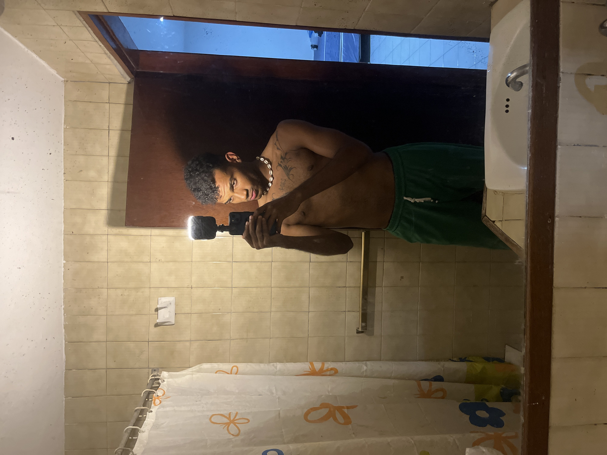 The image shows a mirror selfie taken in a bathroom with light yellow tiles. The subject is a shirtless Black man with short curly hair, a shell necklace, and green shorts. He is holding a camera with a flash attachment. A shower curtain with a floral pattern hangs below. The mirror reflects a view of a window and what appears to be an outdoor scene. While it is impossible to confirm the exact location in Porlamar, Venezuela from this image alone, the bathroom's style and the outdoor view could potentially align with residential or hotel settings in that city.