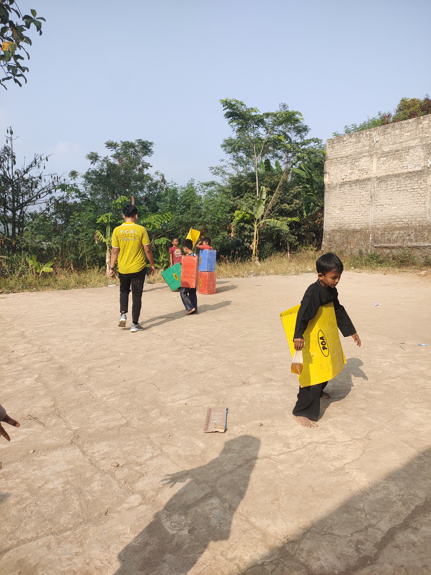 In Subang, Indonesia, on a dirt ground under a clear blue sky, a group of children and an adult are gathered. The adult, wearing a yellow t-shirt with text that reads "PGRI SUBANG," walks away from the camera. In front of him, three children are engaged in an activity. One child, wearing a yellow apron and black pants, carries a yellow box labeled "POA" and a clear plastic bag with a yellowish liquid. To his left, two other children are handling large, colorful cardboard boxes stacked vertically. One box is green, another red, and a third is blue. Behind them, lush green trees and vegetation border the scene, with a textured, light-colored building in the background. The ground is a dusty, sandy earth with patches of dry grass and scattered debris, indicating a semi-rural or outdoor recreational area. The shadows cast by the people and trees suggest it is daytime, likely late morning or early afternoon.